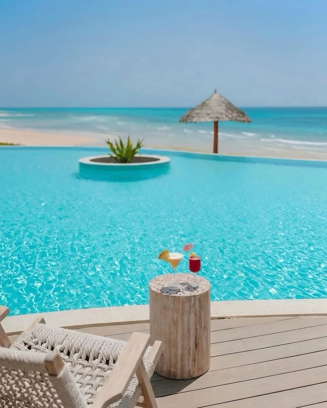 Poolside view with two drinks on a wooden table, pool, beach, and ocean in background, thatched umbrella, sky