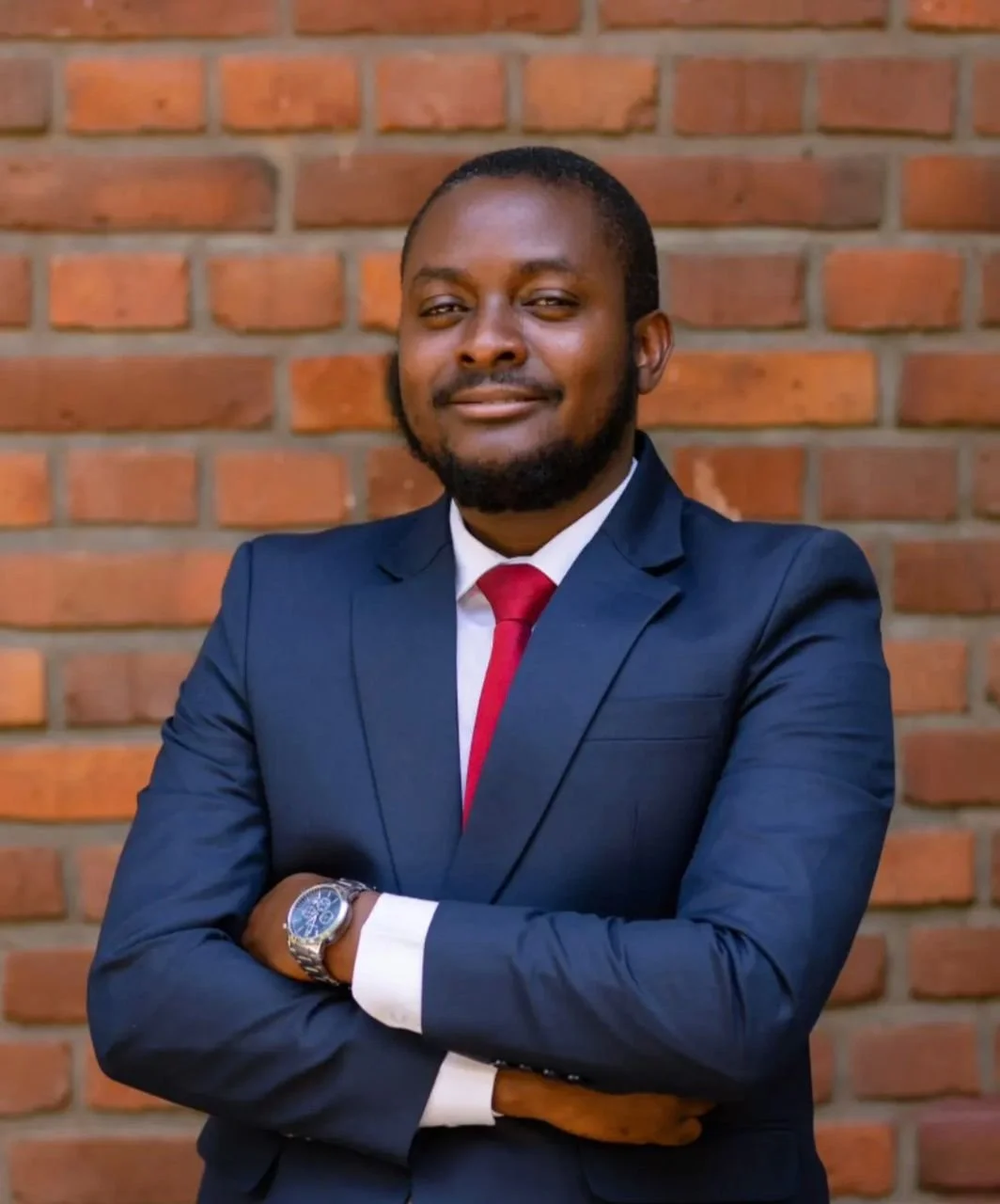 A man dressed in a dark blue suit, white shirt, and red tie standing with arms crossed in front of a red brick wall.