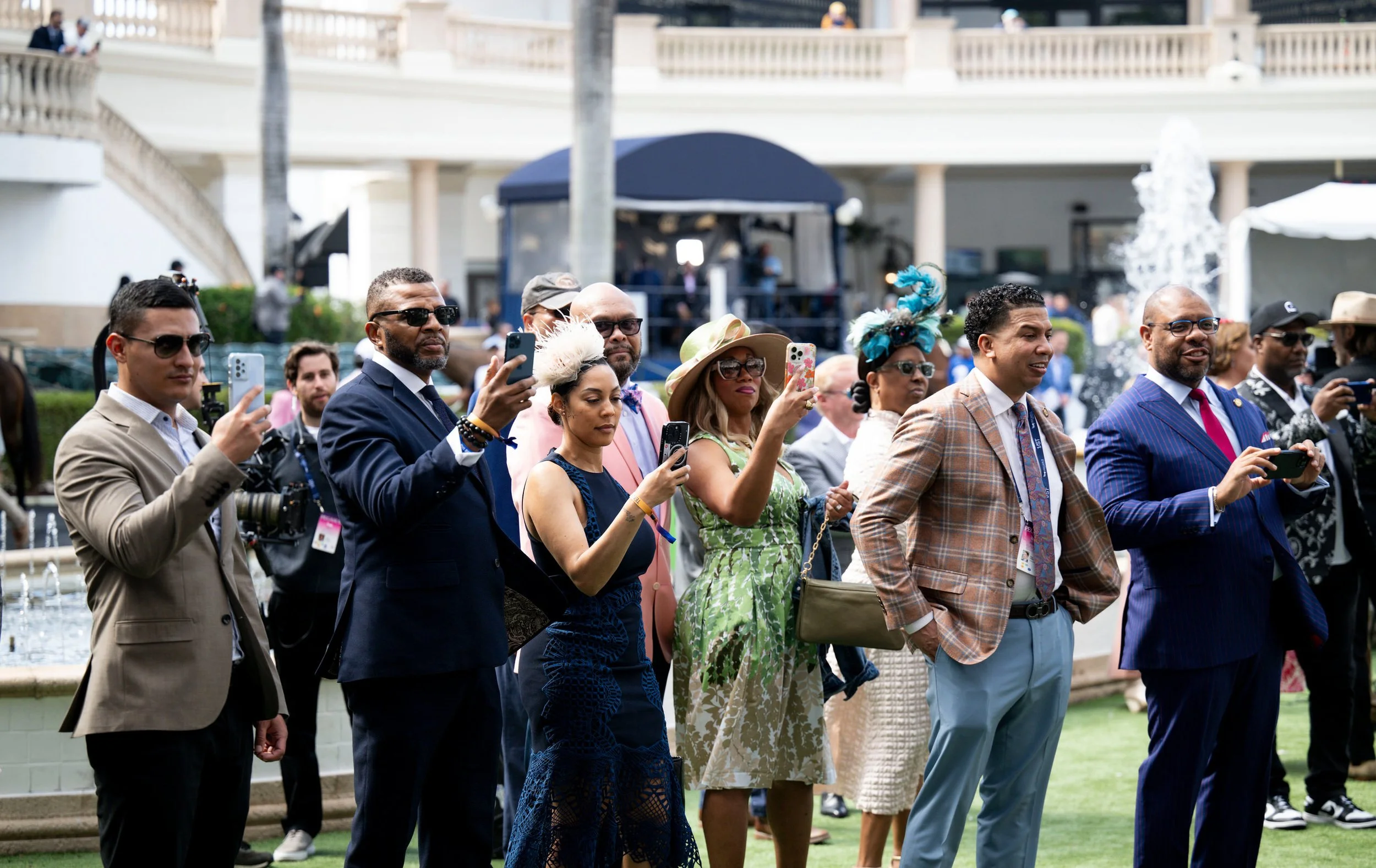 Group of well-dressed people, mostly men wearing suits and women wearing fashionable dresses and hats, standing outdoors and taking pictures with smartphones during a public event or celebration.