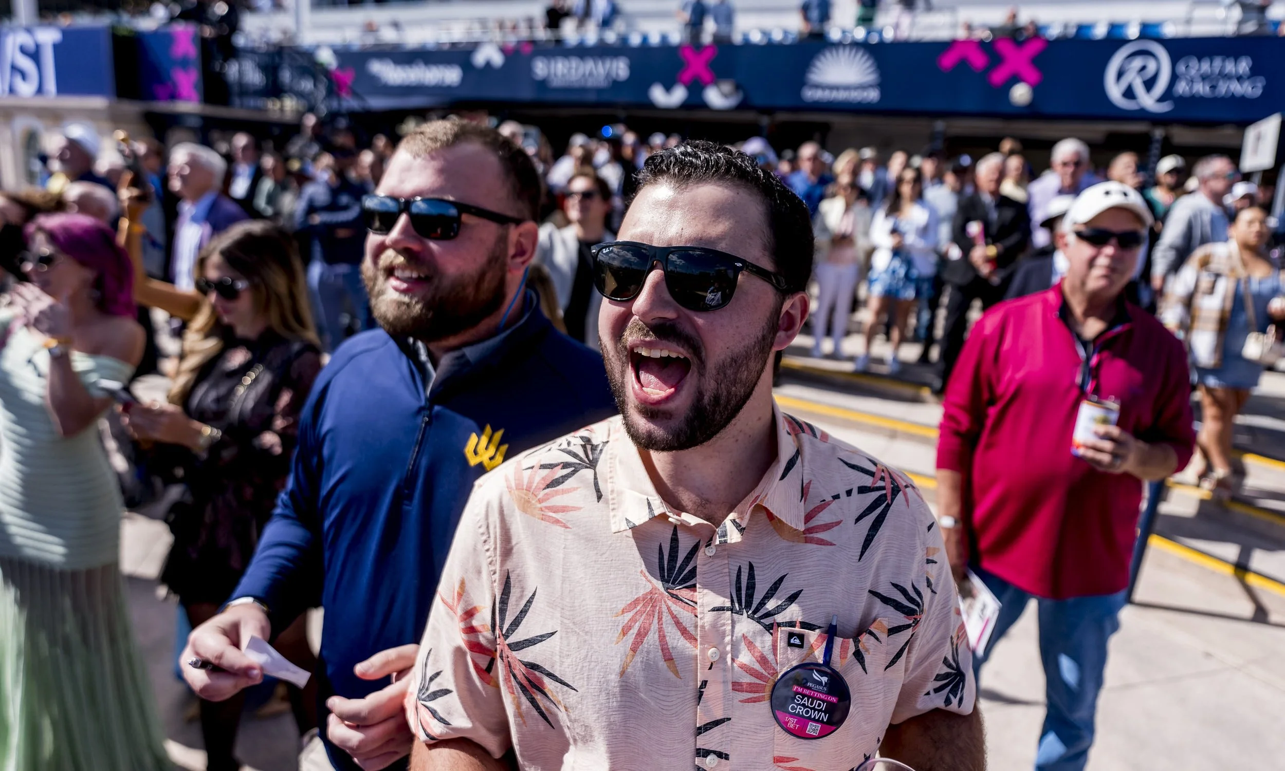 Two men wearing sunglasses, one in a tropical print shirt and the other in a blue jacket, smiling and walking at a crowded outdoor racing event with many people in the background.