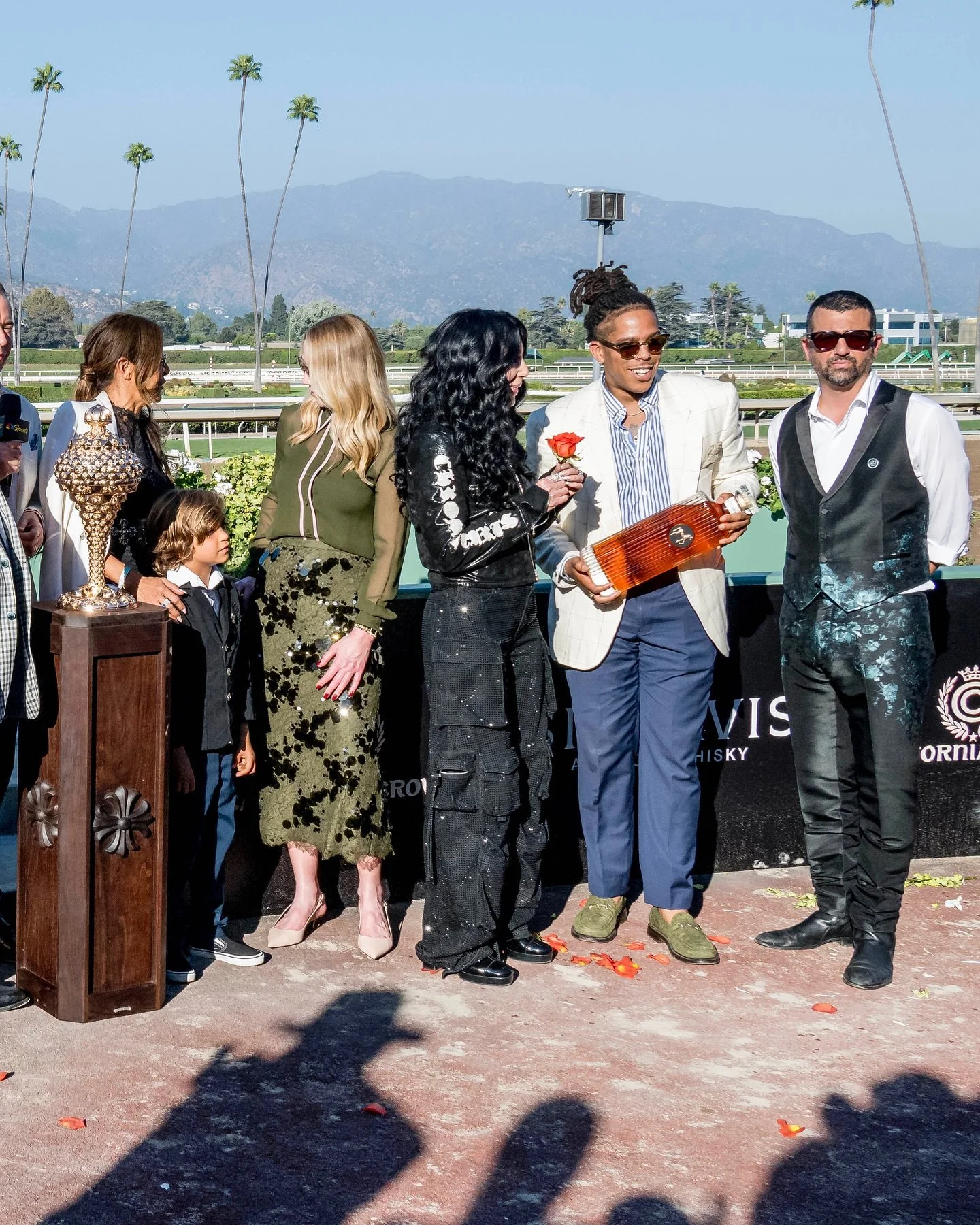 A group of people standing on a racetrack with mountains and palm trees in the background. One person is holding a rose, and another is holding a wooden trophy. There are women and children present, dressed in fancy clothing, and a man in a suit and sunglasses. A podium with a large ornate trophy is visible on the left.