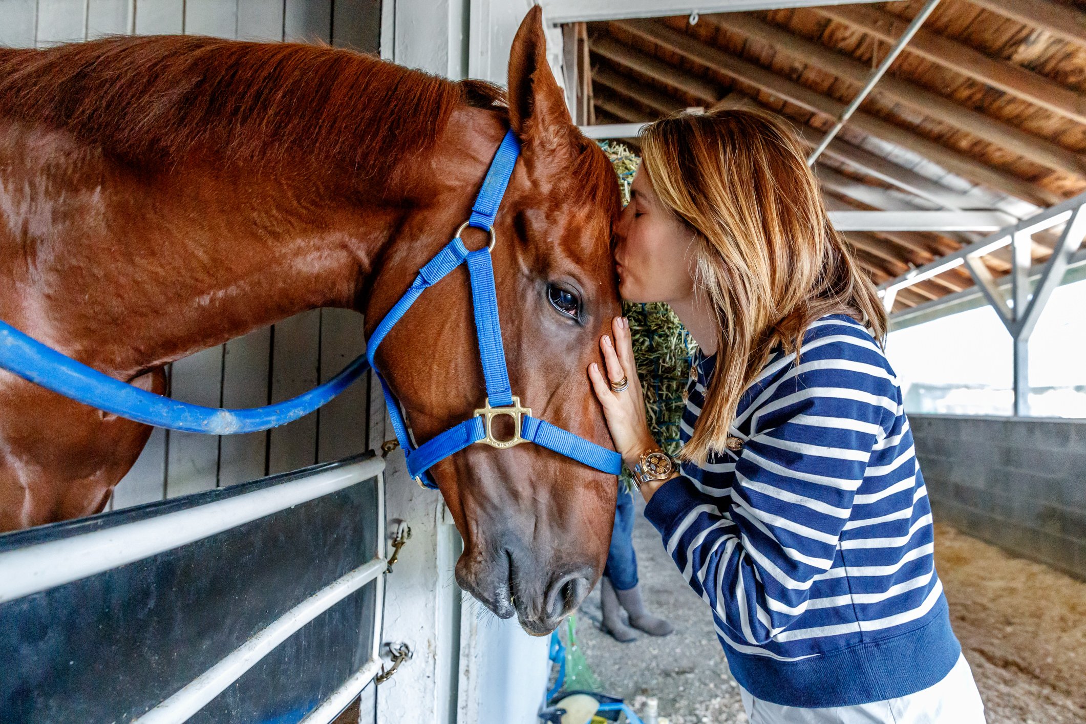A woman kisses a racehorse horse with a blue halter on its head inside a stable.
