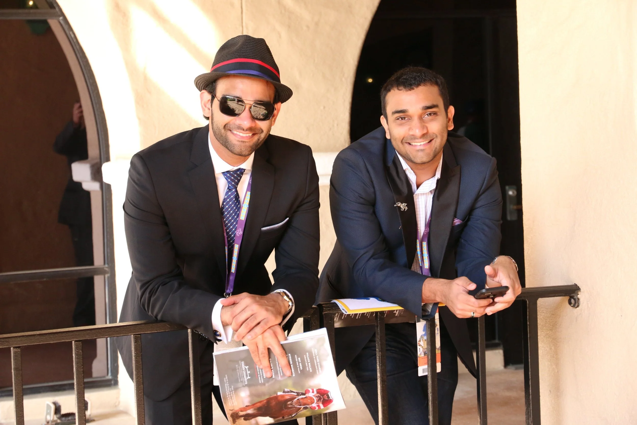 Two men in suits smiling at the camera, one wearing sunglasses and a hat, the other holding a phone, leaning on a railing at a Thoroughbred racing event.
