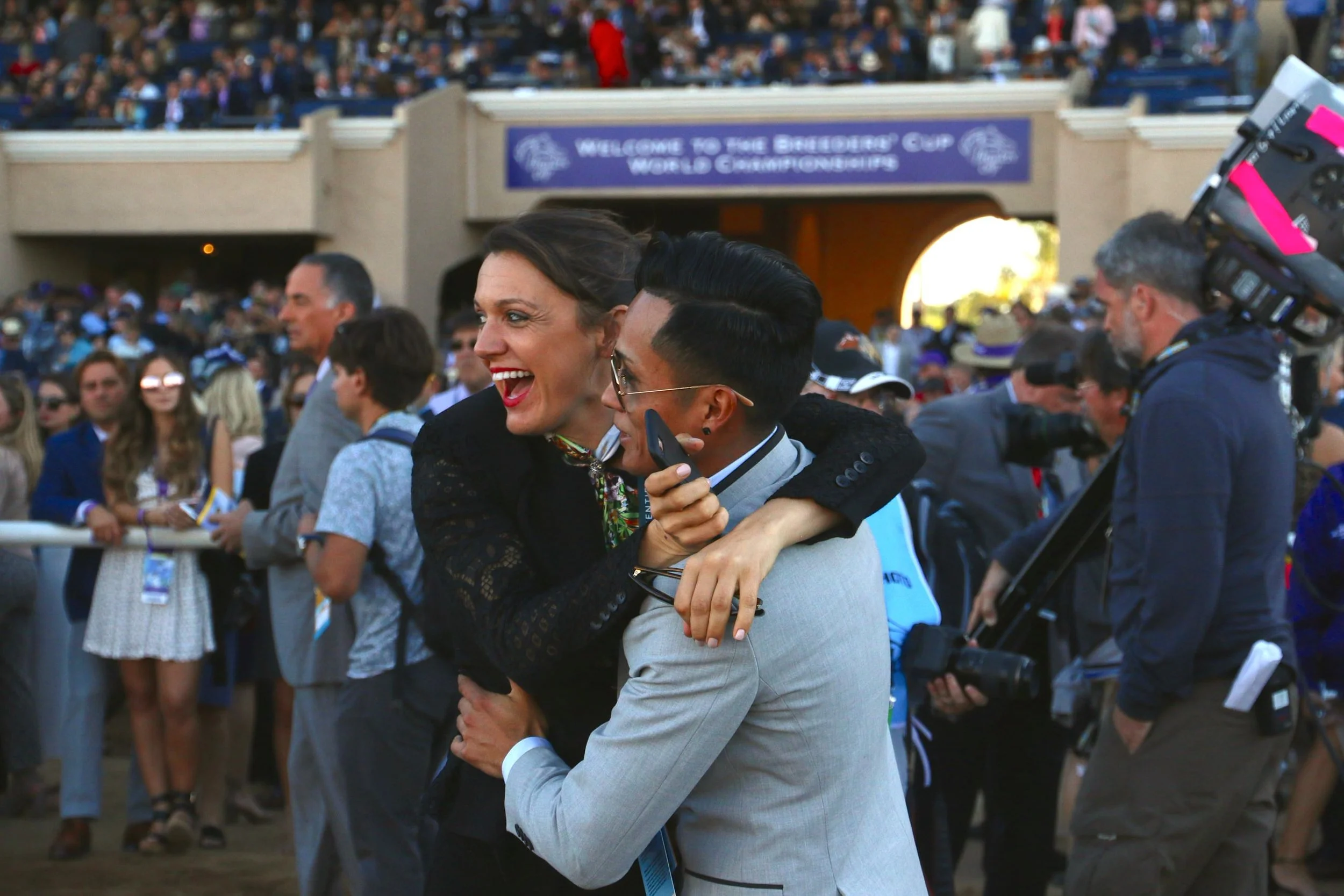 Two people celebrating, hugging at an outdoor Thoroughbred racing event with a crowd behind them, with a sign reading 'Welcome to the Breeders' Cup World Championships' in the background.
