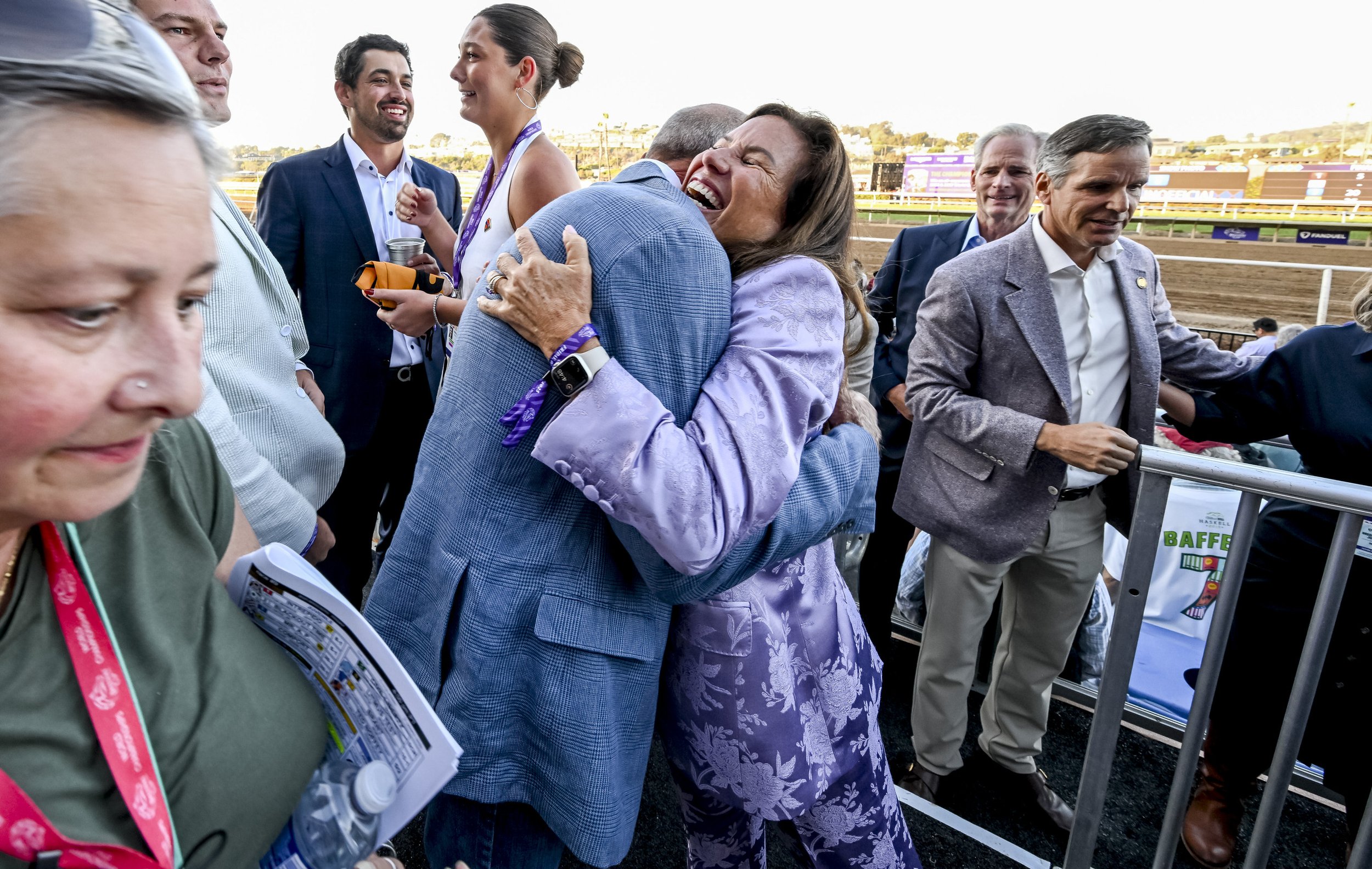 Two women hugging at a racetrack event, surrounded by several men, some smiling and others observing, with a racetrack and colorful advertisements in the background.