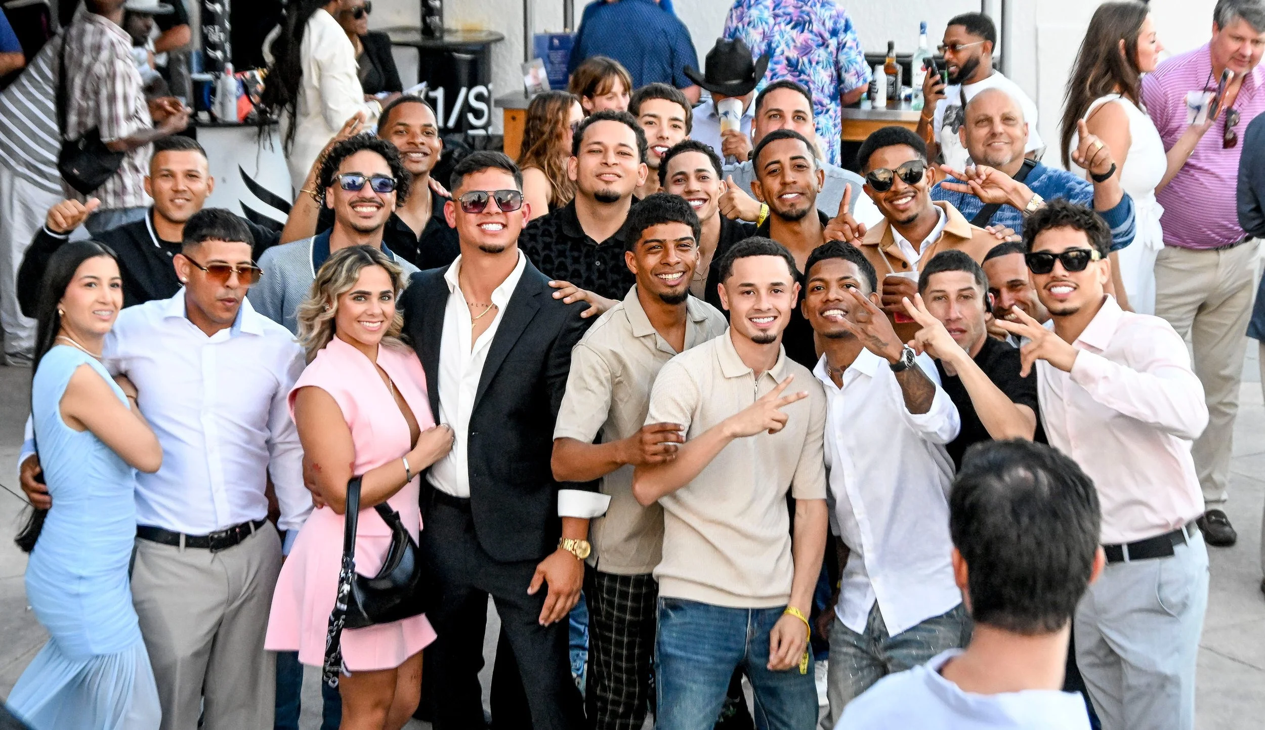 Group of smiling young adults posing together outdoors, some making peace signs, with a diverse mix of men and women dressed in casual and semi-formal clothing, in a social gathering setting at a race track.