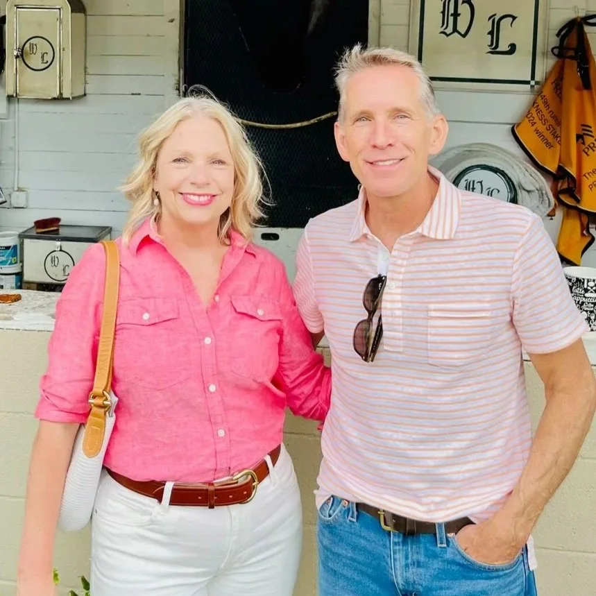 A smiling woman in a pink blouse and white pants standing next to a smiling man in a striped polo shirt and blue jeans, outdoors against a white wall with various items and decorations around.