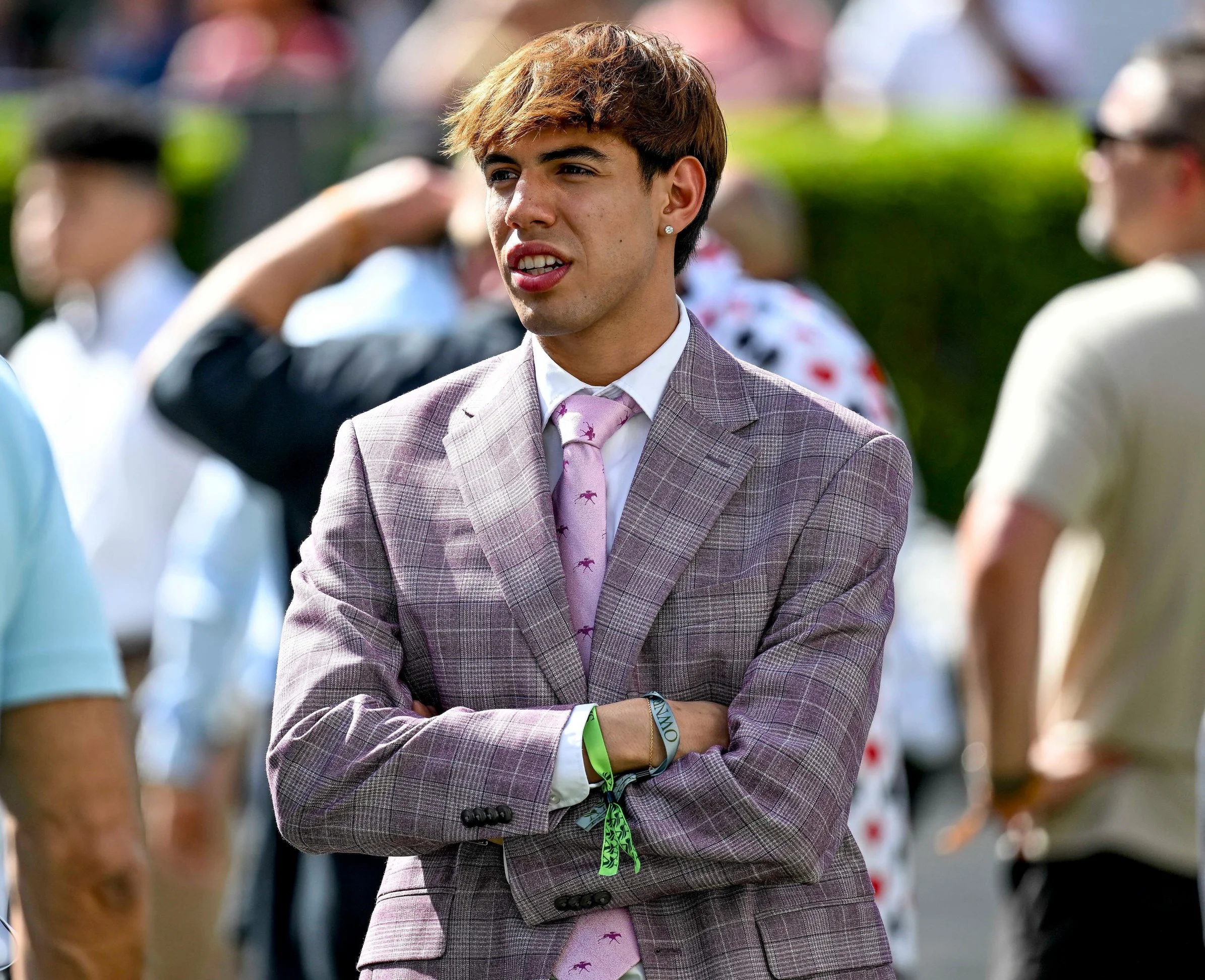 A young man with light brown hair wearing a plaid suit and a pink tie with small horse patterns, standing outdoors at a race track with his arms crossed, surrounded by people.