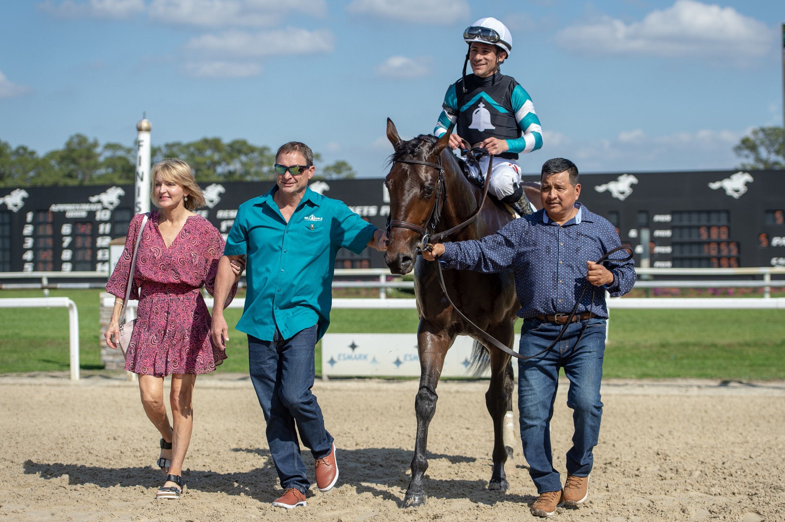 A person in racing attire on a brown horse, accompanied by three people walking on a racetrack with a black scoreboard in the background.