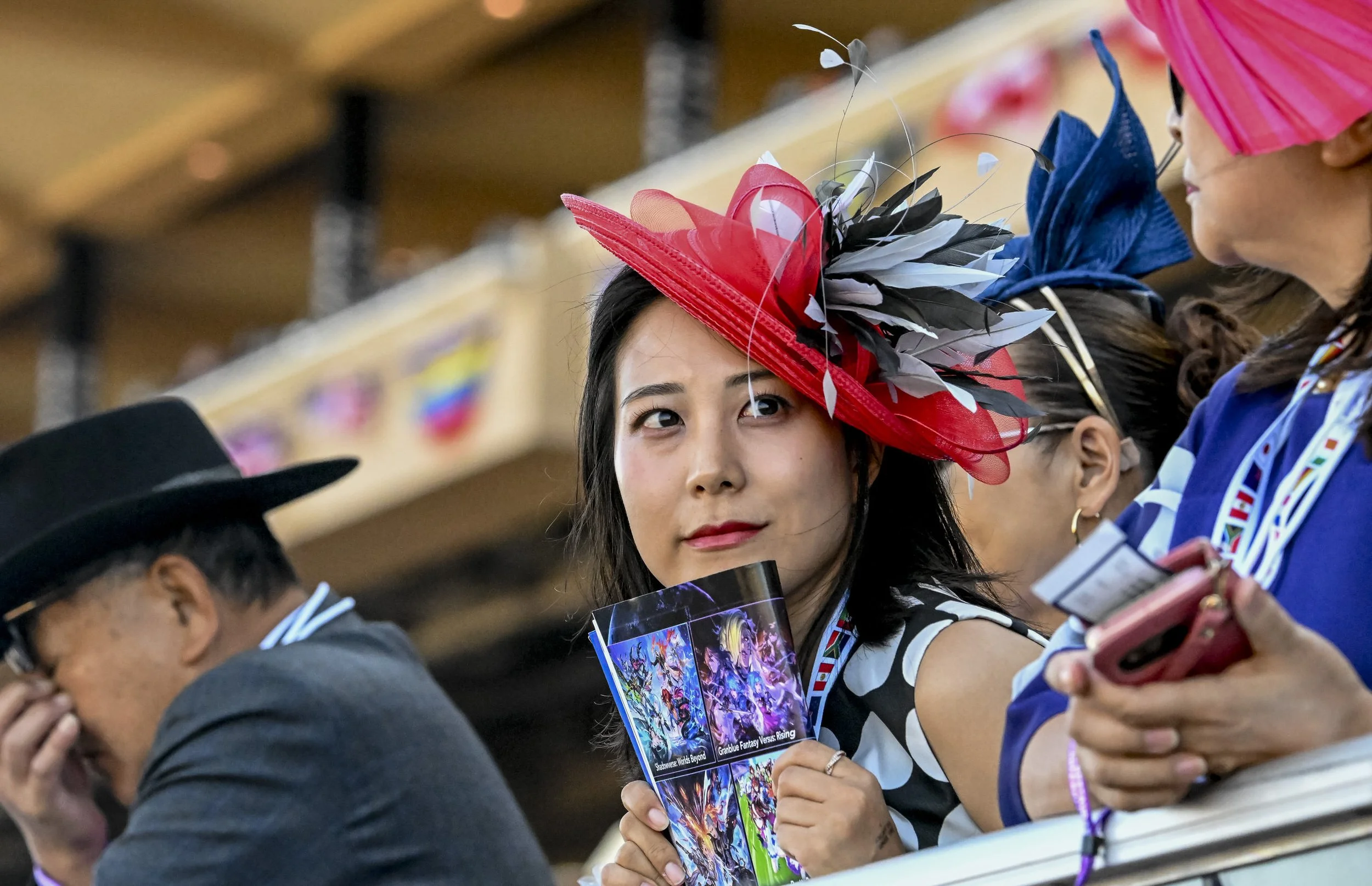 A woman wearing a large red hat with black and gray feathers looks to the side at a parade or event, holding a fan with colorful artwork. Other women nearby are wearing elaborate hats and are engaged with their phones or talking.