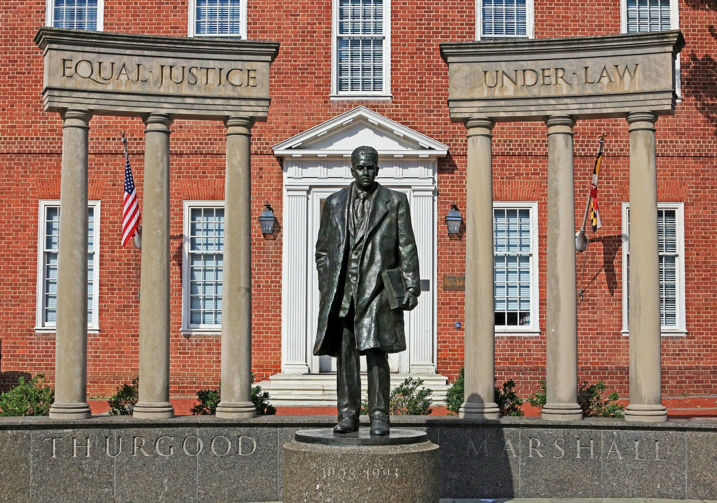 Statue of Thurgood Marshall in front of a brick building with six windows and a sign that reads "EQUAL JUSTICE UNDER LAW." The statue depicts Thurgood Marshall holding a book, wearing a suit and overcoat. The base of the statue has his name and lifespan, "1968-1993."