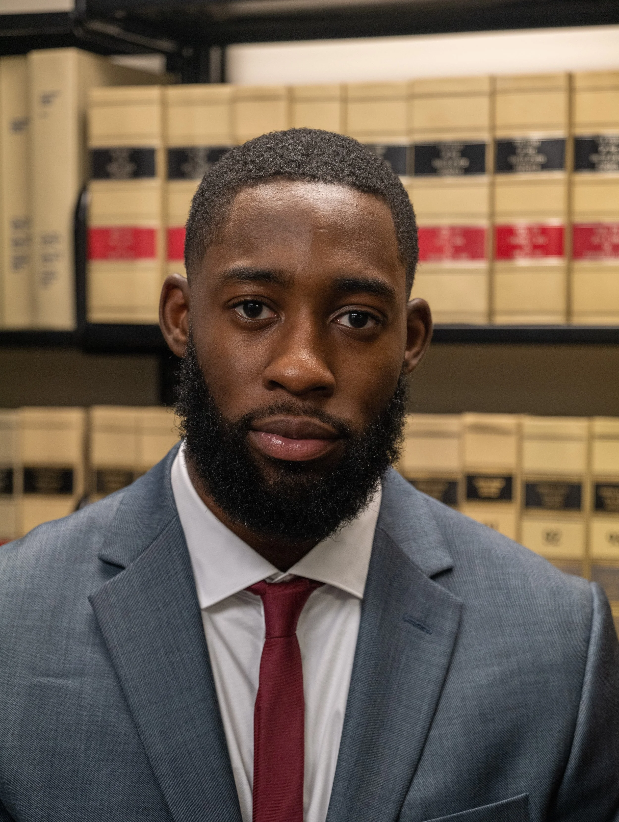 A man in a gray suit, white shirt, and red tie standing in front of shelves with legal books.