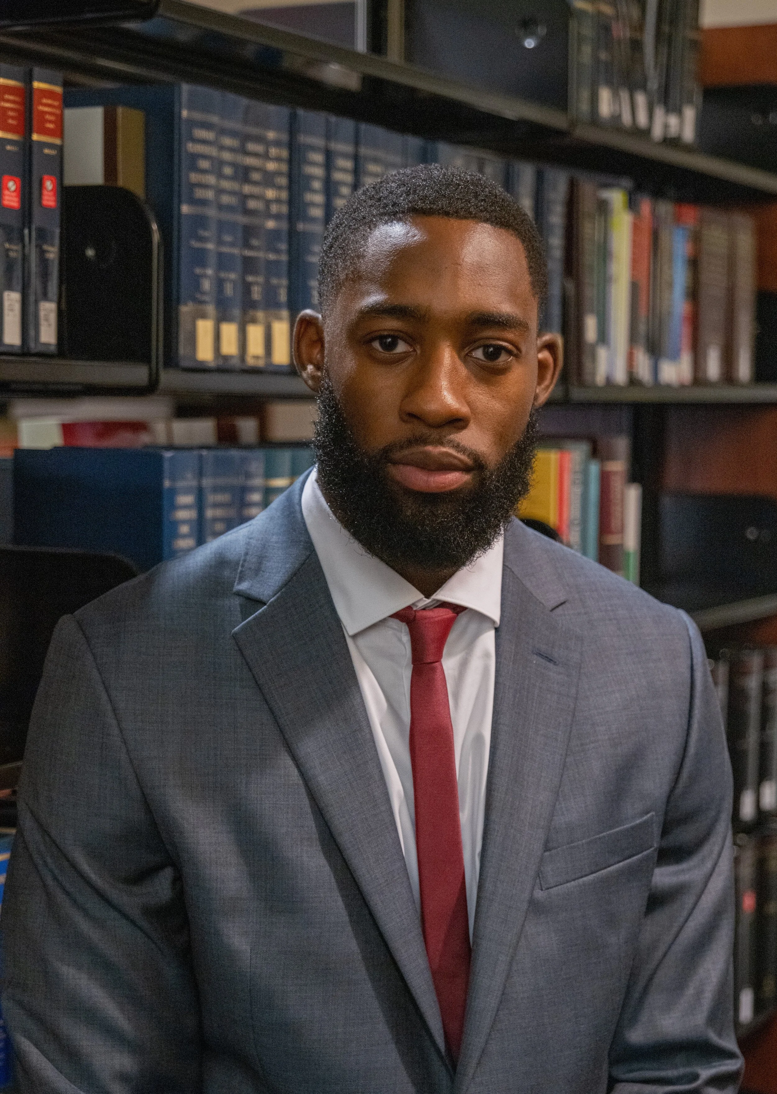 A young man in a gray suit and red tie sitting in a library with shelves of books behind him.