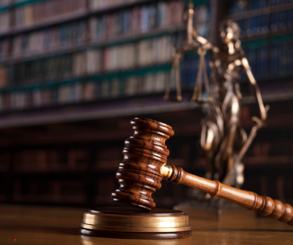 A judge's wooden gavel resting on a sound block, with a blurred statue of Lady Justice holding scales in the background, in a law library setting.