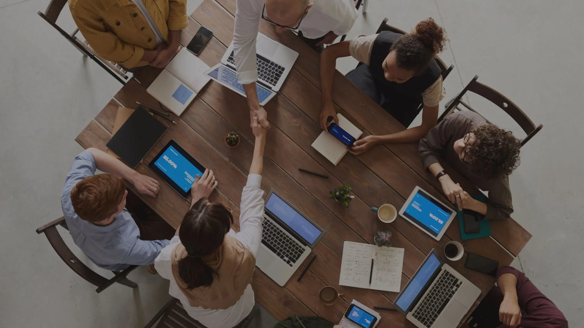 A top-down view of seven people sitting and standing around a wooden conference table with laptops, tablets, notebooks, pens, and coffee cups during a business meeting.