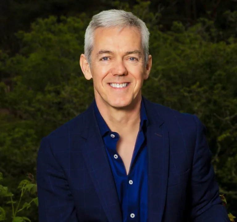 A smiling middle-aged man with gray hair wearing a dark blue blazer and matching shirt, standing outdoors with green foliage in the background.