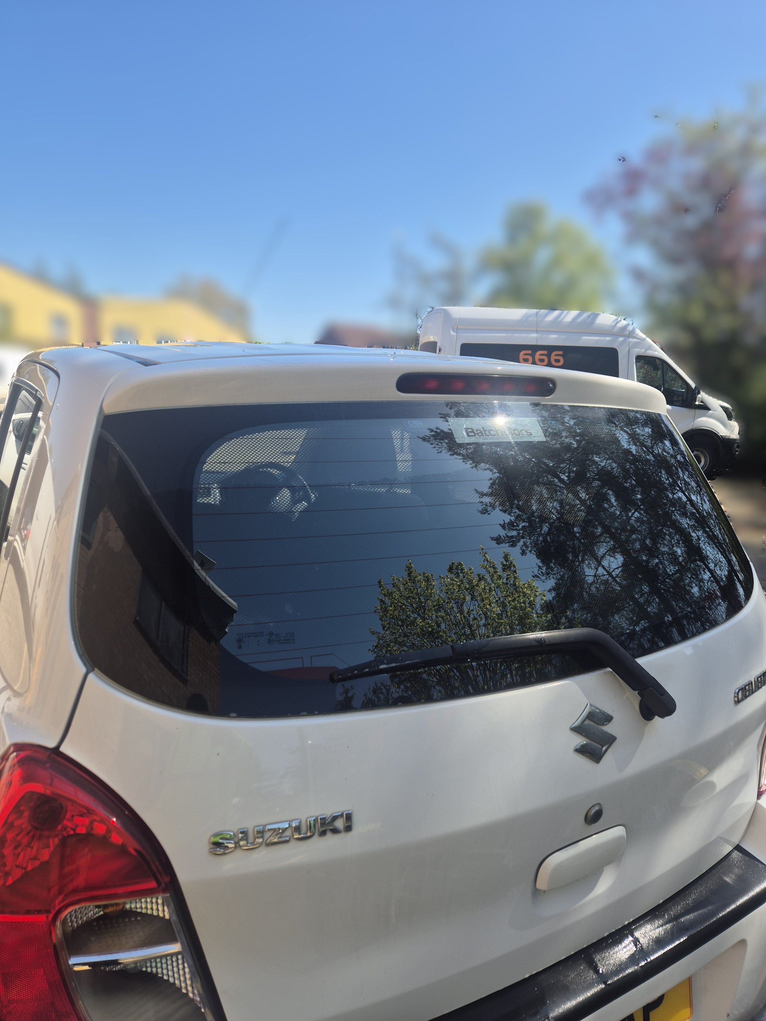 Rear view of a white Suzuki vehicle with tinted windows and a red taillight, parked outdoors with trees and buildings in the blurred background, under clear blue sky.