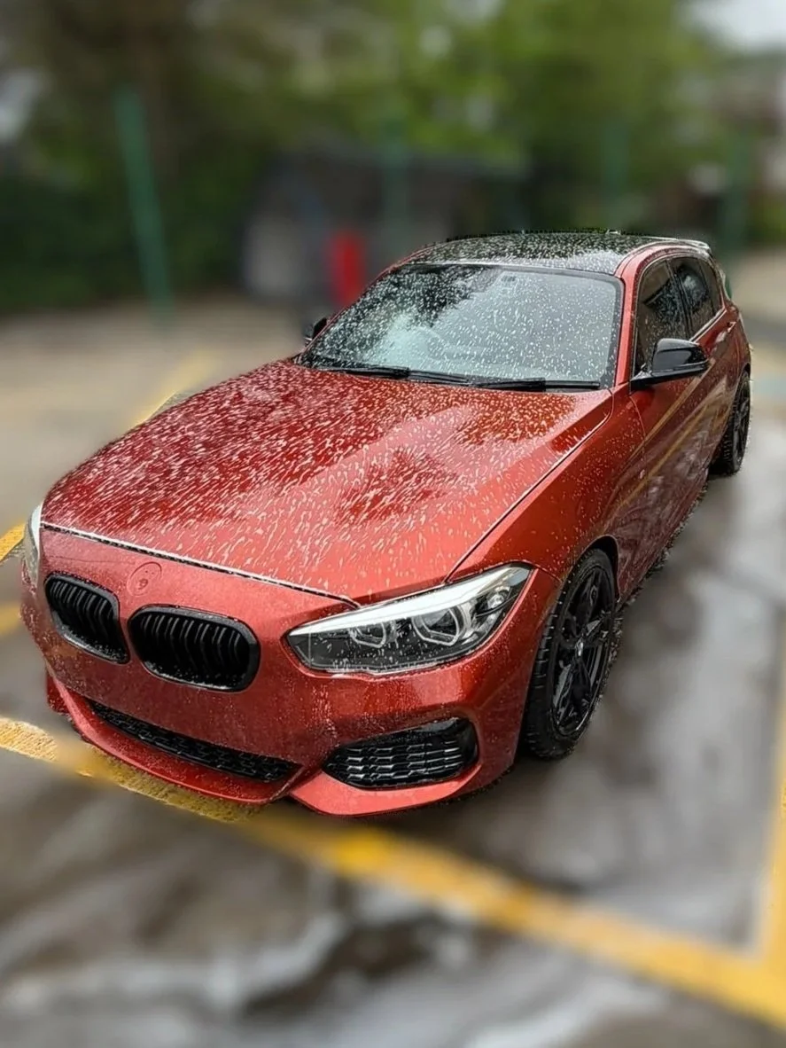 Red sport car with black wheels parked on a wet surface, covered in rain water droplets, with a blurred green background.