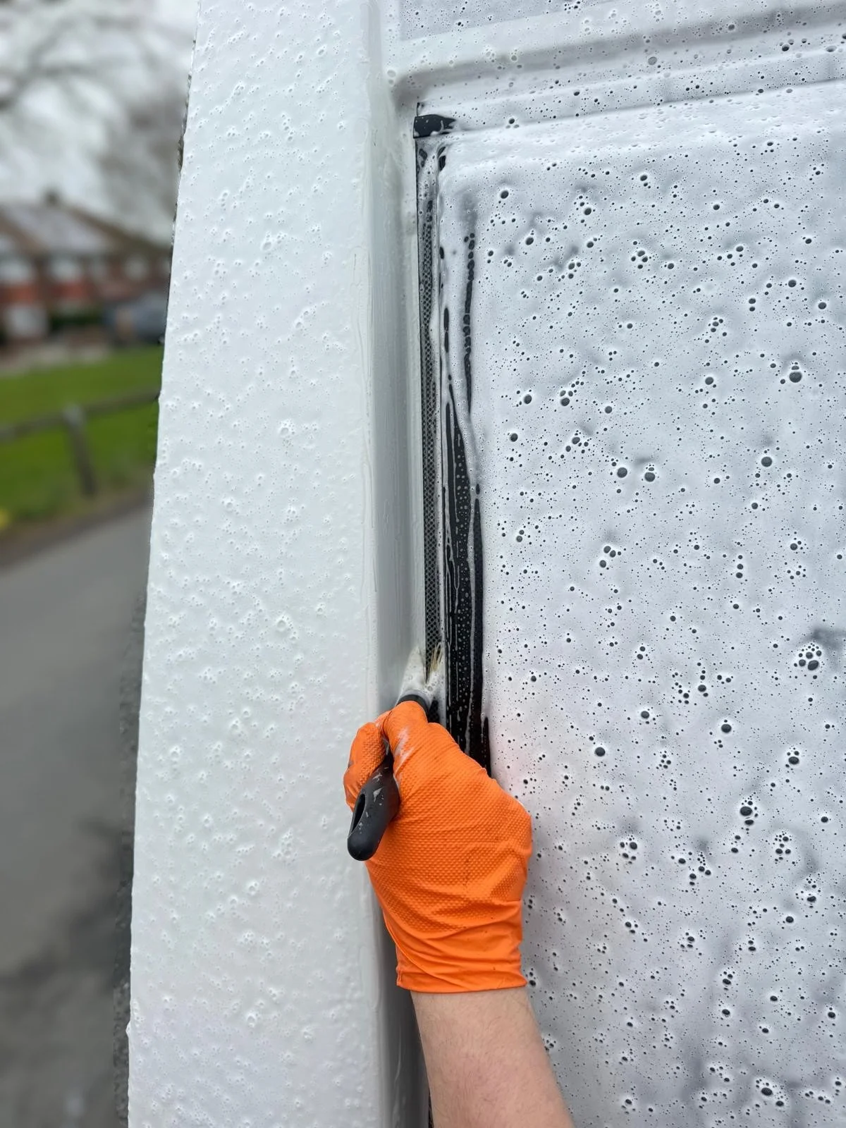 A person wearing an orange glove using a cleaning brush to remove dirt from the edge of a white caravan surface outside, with soap bubbles on the foam.