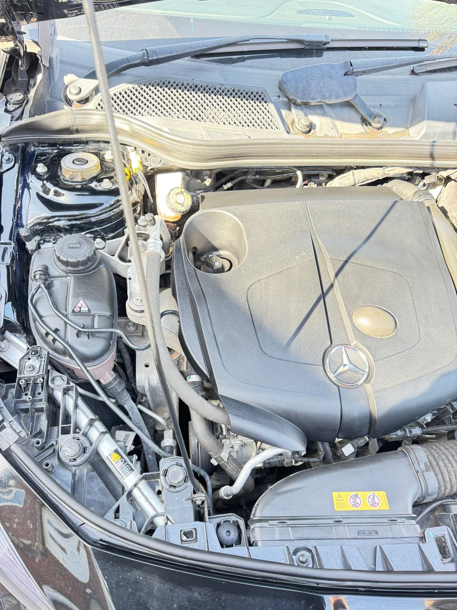 The image shows the engine bay of a Mercedes-Benz vehicle, with various engine components, hoses, and compartments visible.