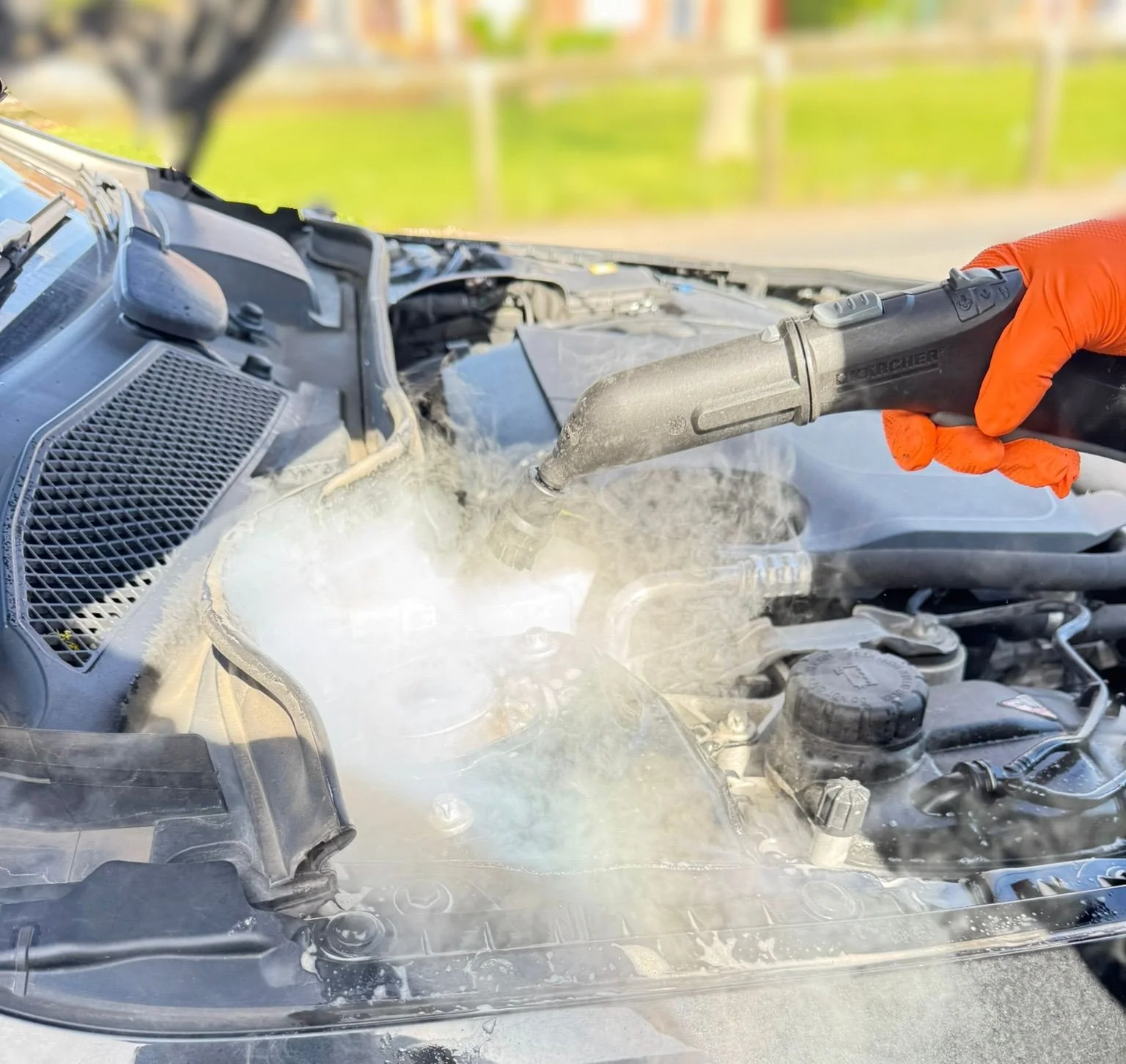 A person wearing orange gloves cleaning the engine bay of a car with a steam cleaner.