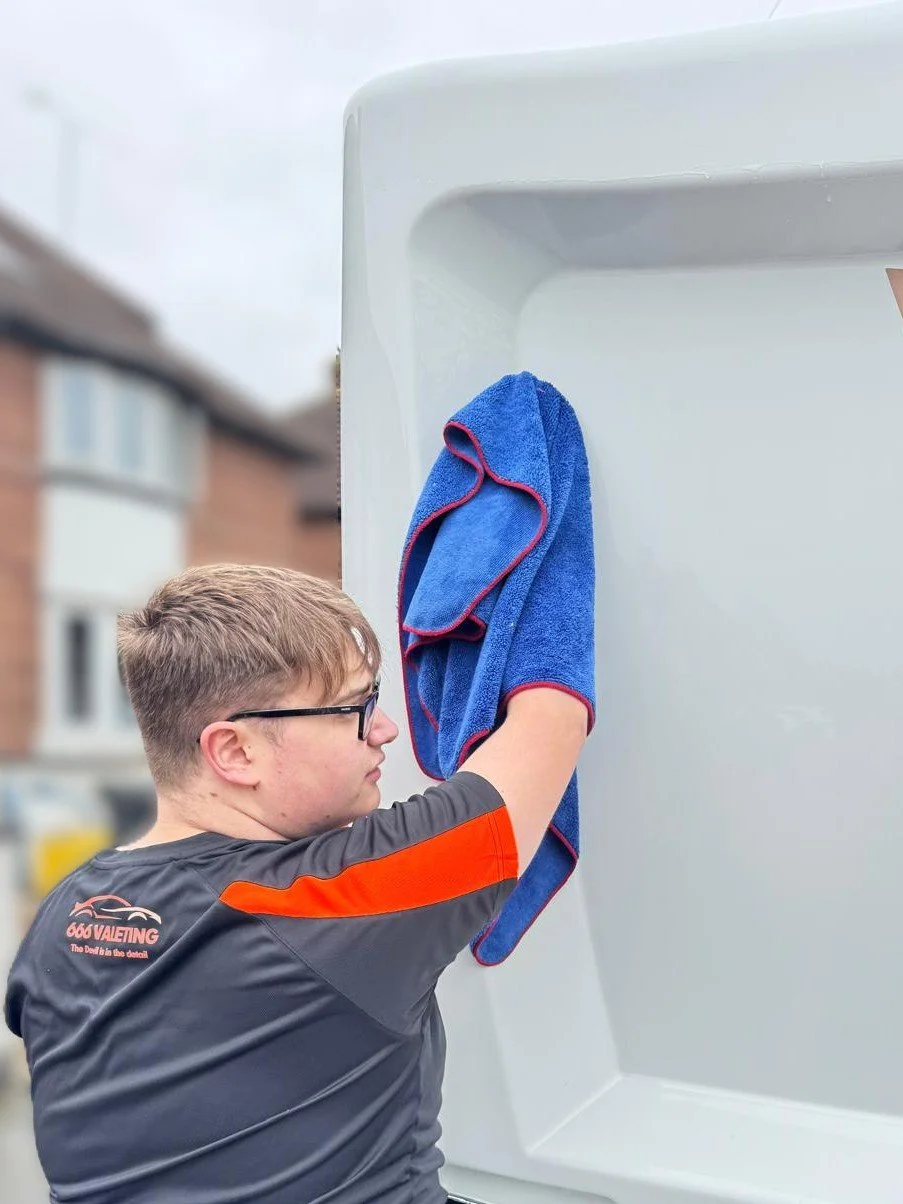 A young man with glasses in a black shirt with red accents, cleaning the outside of a white caravan with a blue towel.