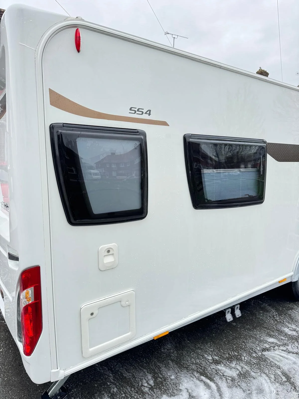 A clean white camper trailer with two black-framed windows, a small door, a red tail light, and a black wheel on a wet asphalt surface.