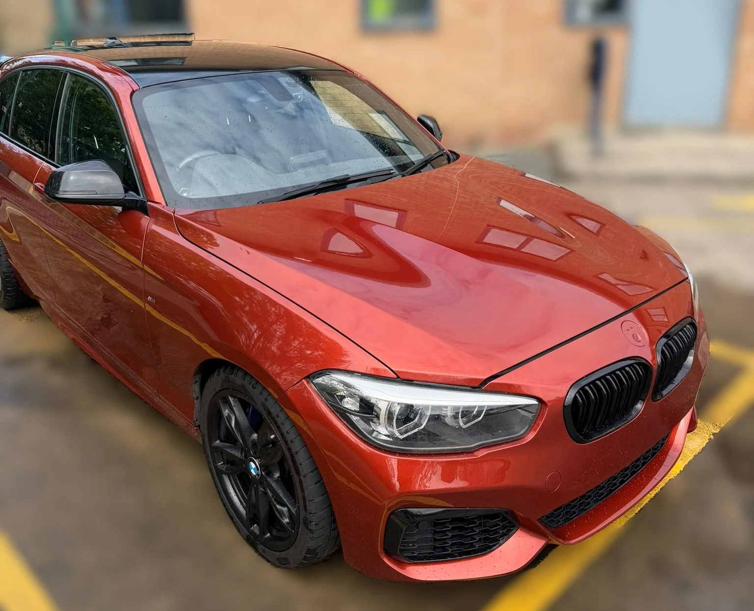 Red BMW car parked in a parking lot near a brick building with windows.