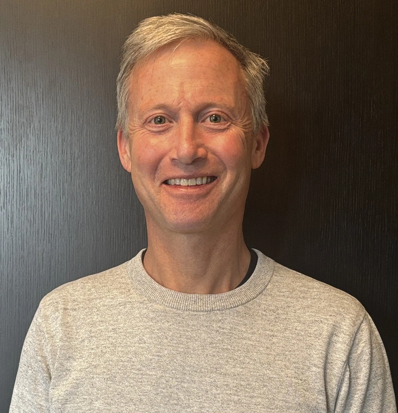 A smiling middle-aged man with gray hair, wearing a beige sweater, standing in front of a dark wooden background.
