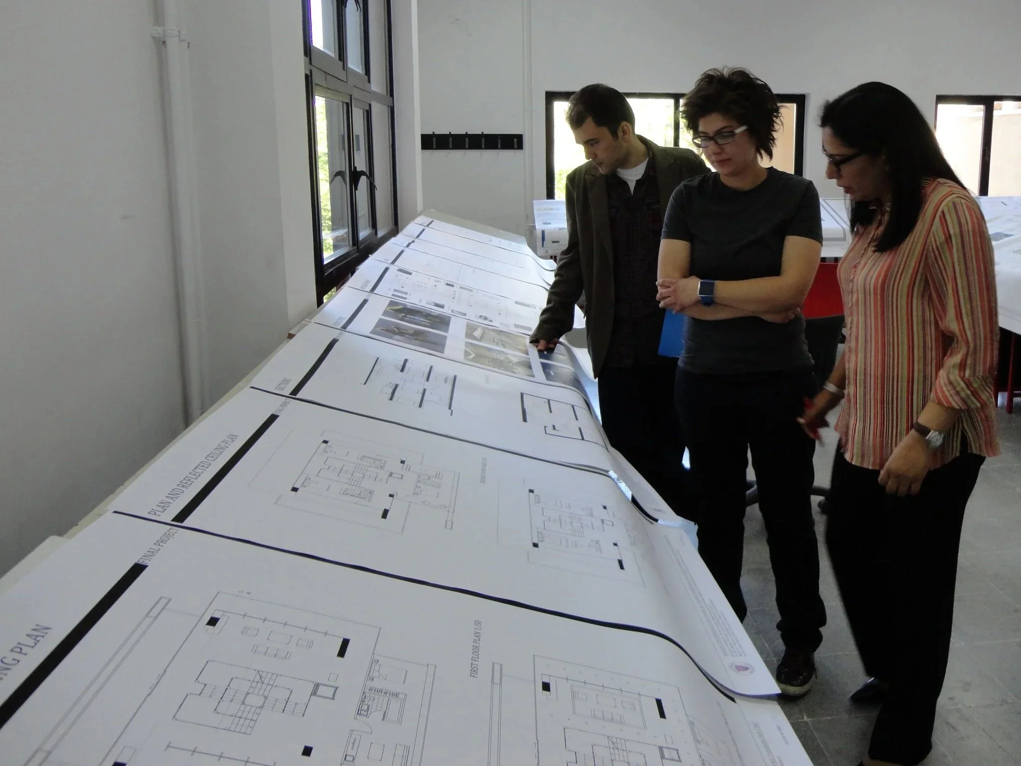 Three people examining architectural blueprints or plans laid out on tables in a bright room with windows.