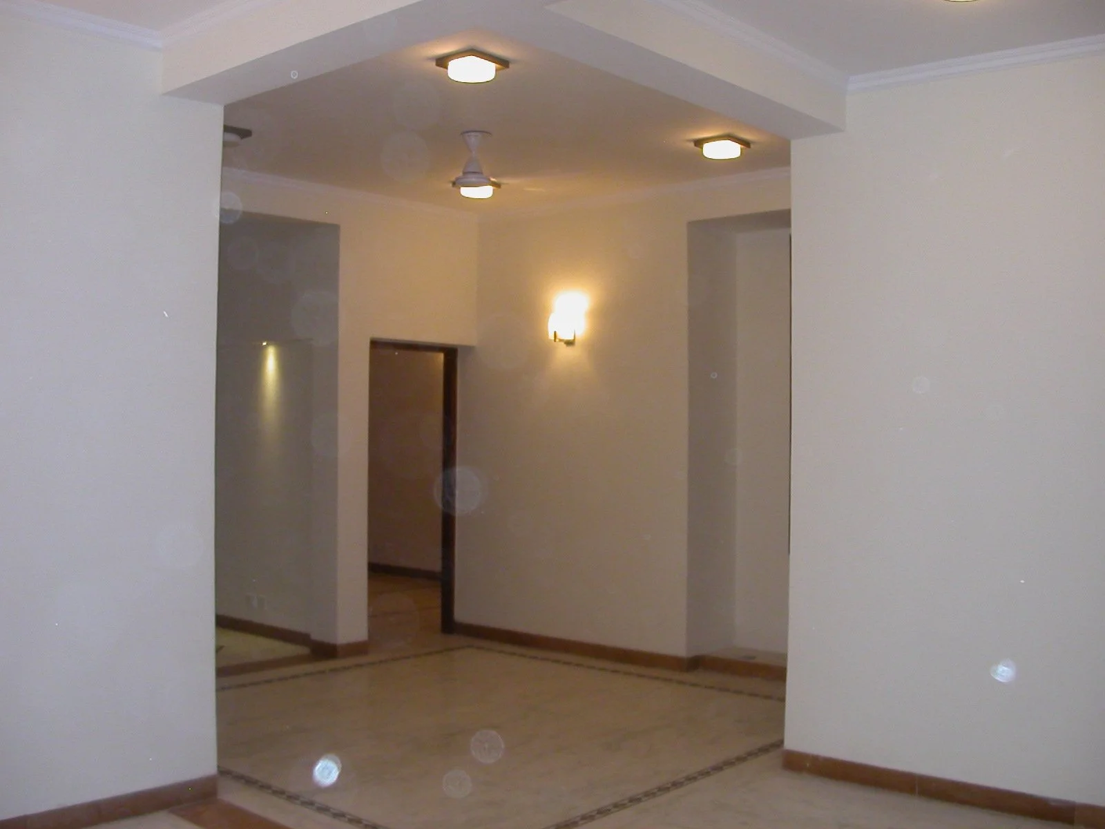 Interior view of a hallway with beige walls, wooden baseboards, ceiling lights, and a partially open door leading to another room.