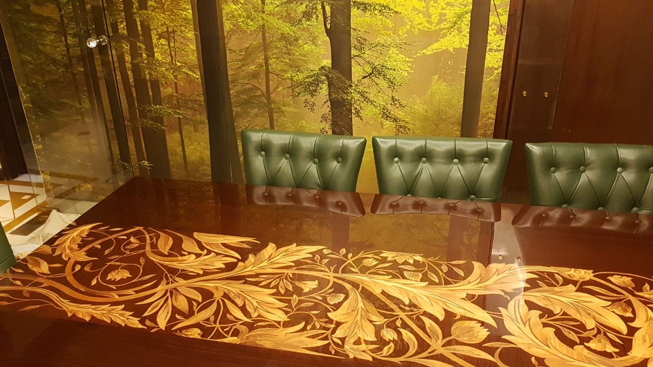 A conference room with a large wooden table featuring intricate gold floral patterns. Around the table are green leather chairs with button tufting. The backdrop is a wall mural of a forest scene with trees and yellow-green leaves.