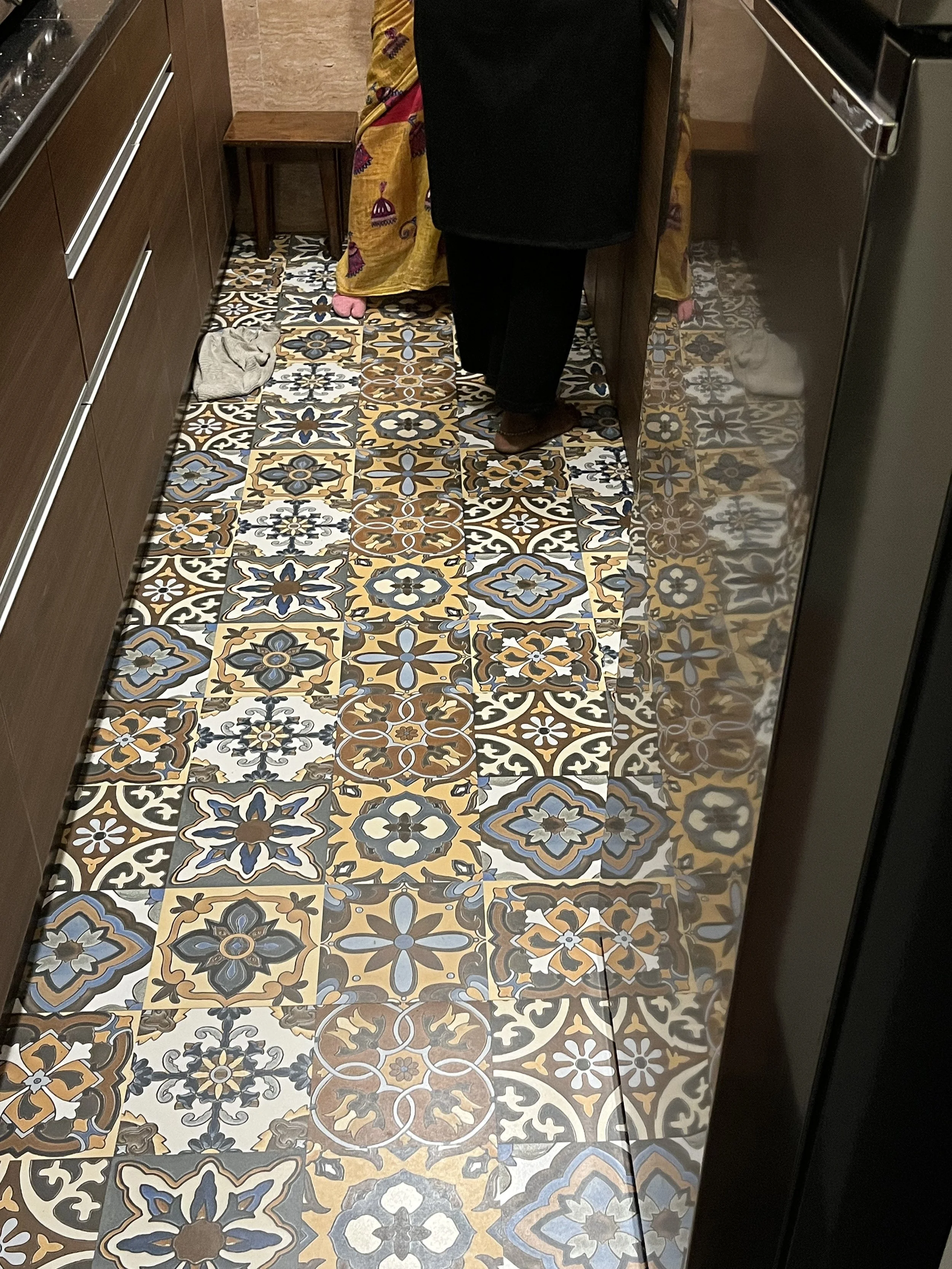 View of a woman in a black coat and pants in a kitchen taking a photo of the floor with patterned tiles. The tiles have a colorful, intricate geometric pattern in shades of yellow, blue, black, white, and brown. There is also a pair of pink slippers at the far end of the room, a small wooden stool, and a towel on the floor.