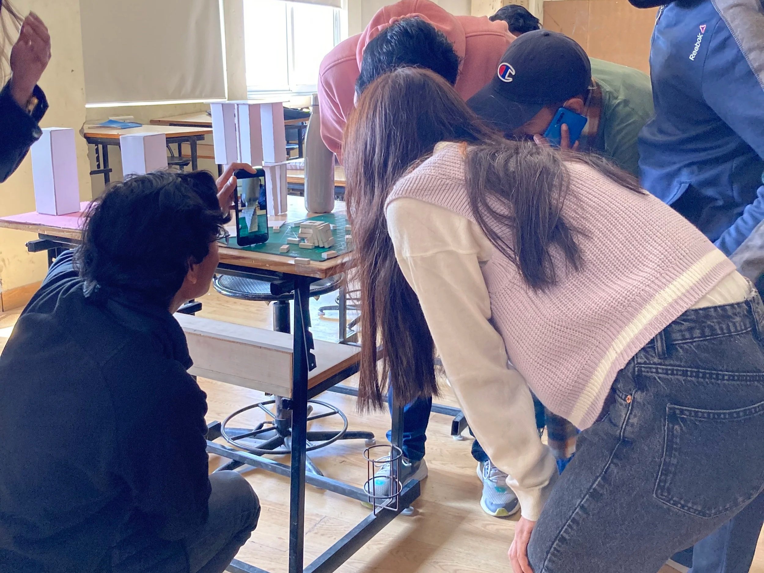 A group of people gathered around a table, observing small white objects, with some individuals taking pictures using their phones. The activity appears to be collaborative or educational, taking place in a bright room with wooden floors and chairs in the background.