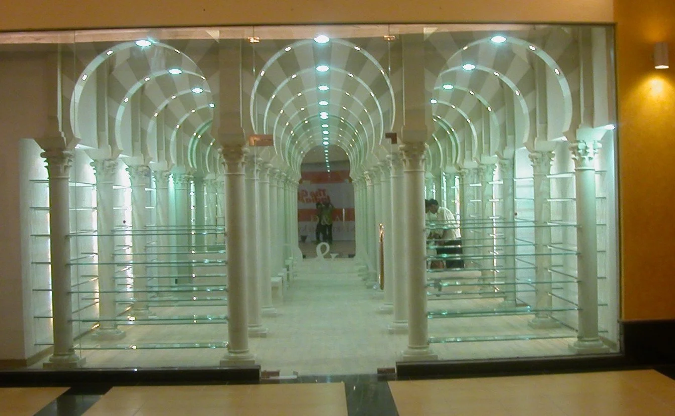A glass wall with an interior mirror installation that creates an illusion of a long corridor with white columns and arches, reflecting ceiling lights.