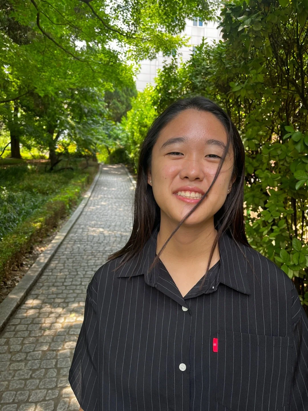 A young woman with long dark hair standing on a cobblestone path surrounded by green trees and bushes, smiling.