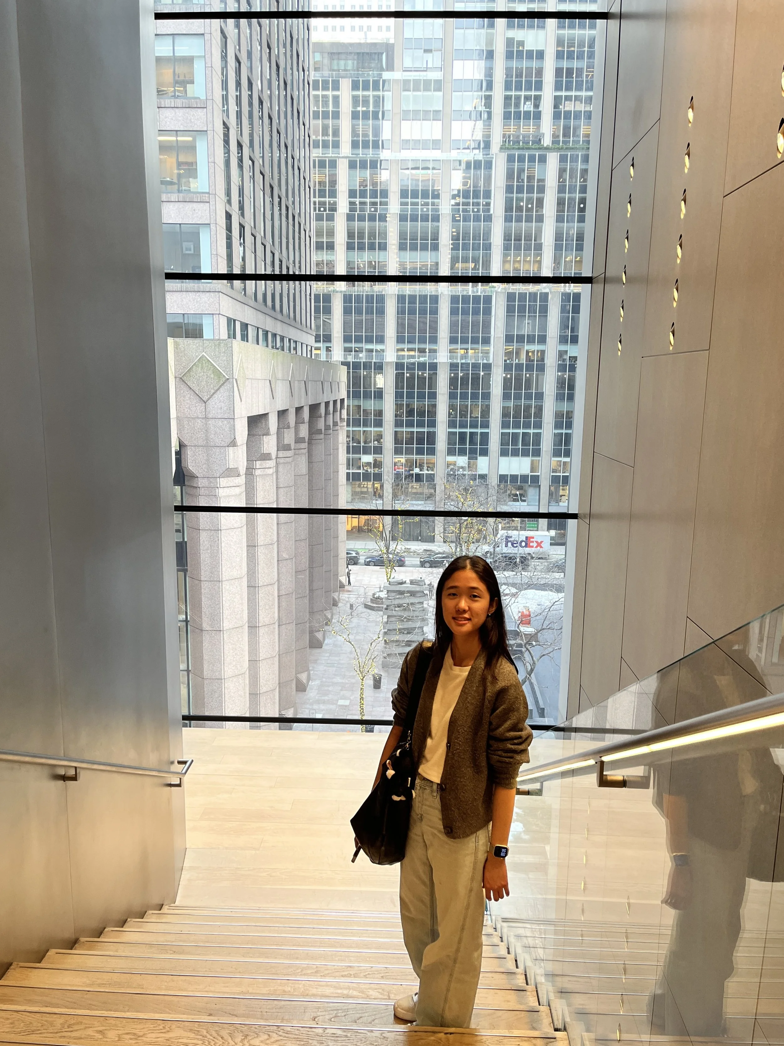 Young woman standing at the top of a staircase in a modern building with large floor-to-ceiling windows overlooking city office buildings.