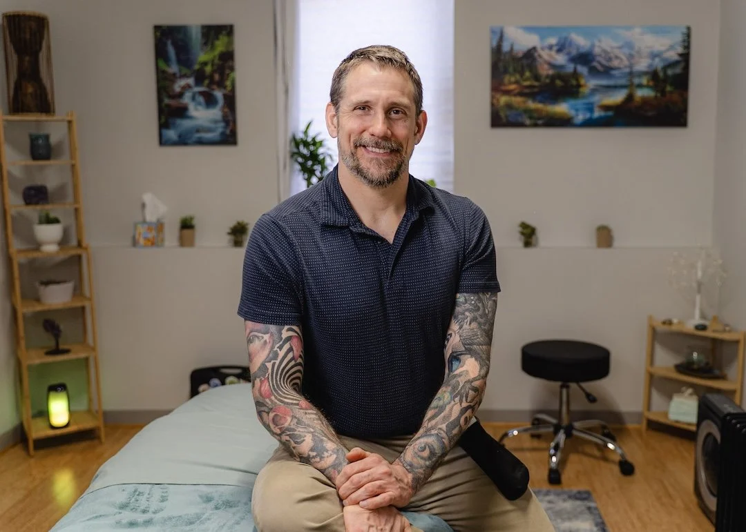 Smiling massage therapist with tattoos on his arms, sitting on a massage table in a room decorated with nature-themed paintings and potted plants.