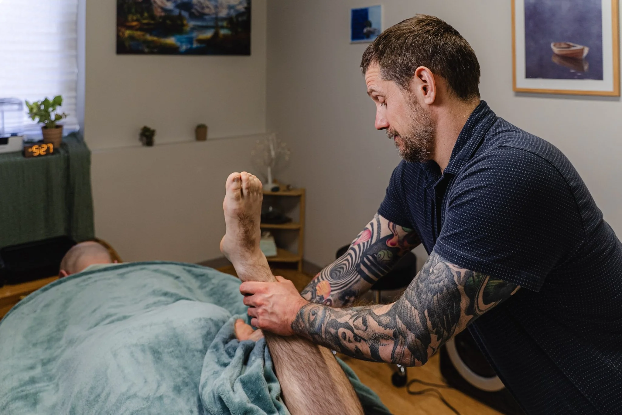 Massage therapist with tattoos stretches right leg of bald male client with painting of river in background