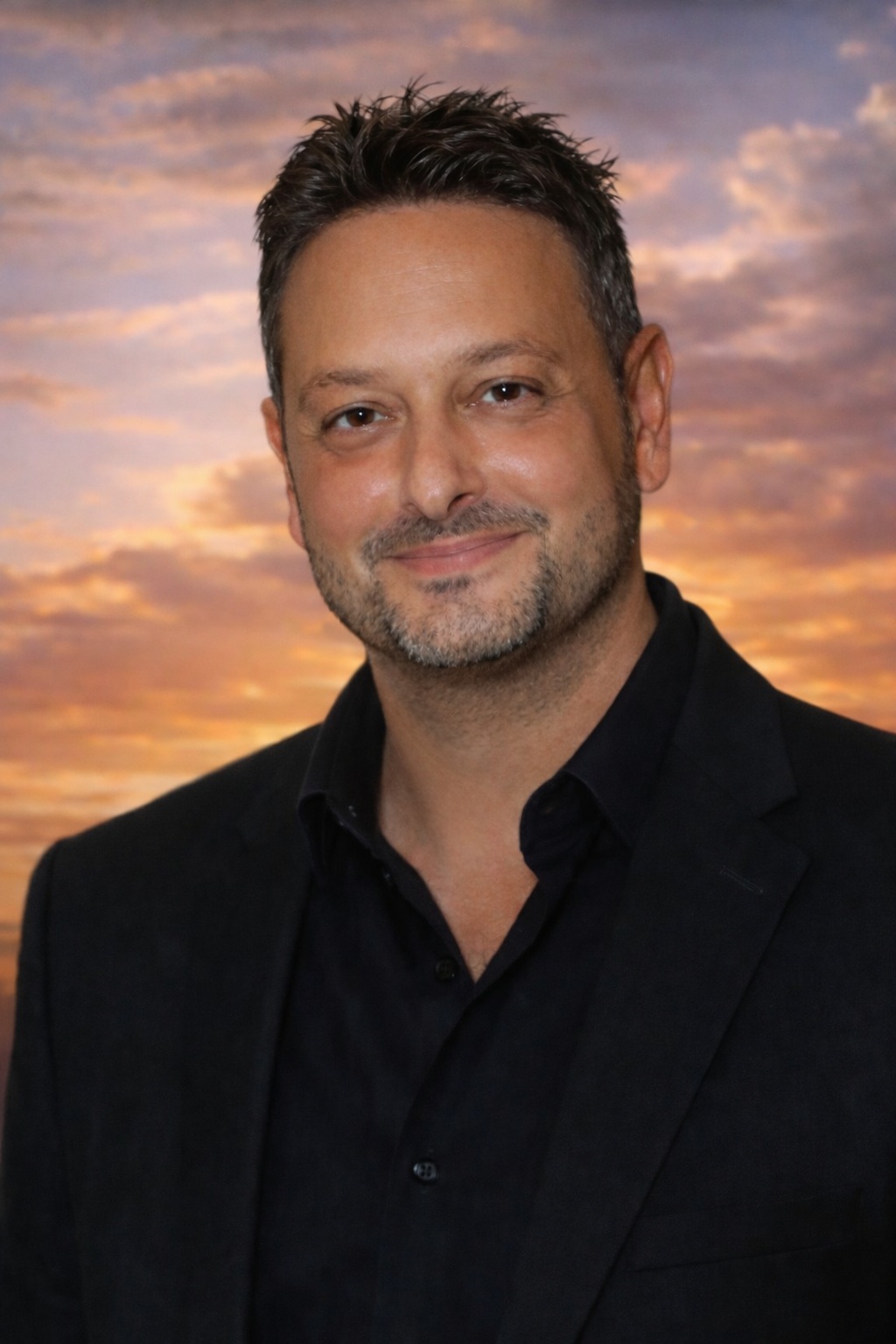 Photograph of a white man with dark hair and a short beard, wearing black with a cloudy sunset in the background