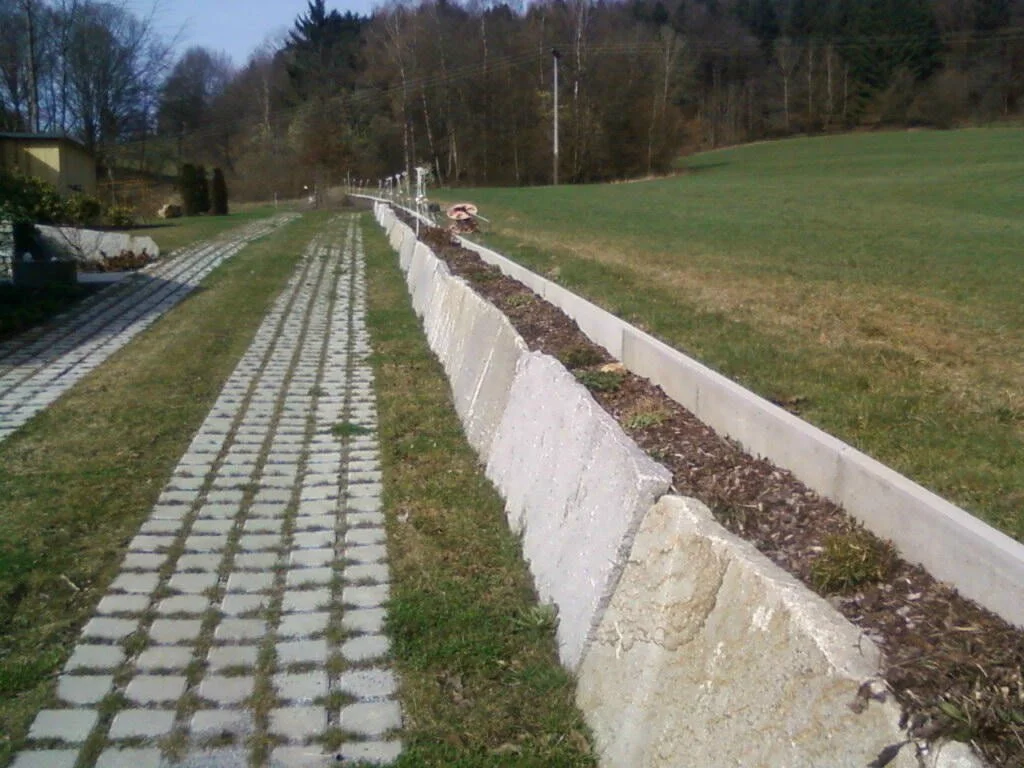 Ein unbefüllter Gehweg mit Pflastersteinen links und einer Baugrube rechts, die durch eine Reihe von Betonrampen und Absperrungen gesichert ist.