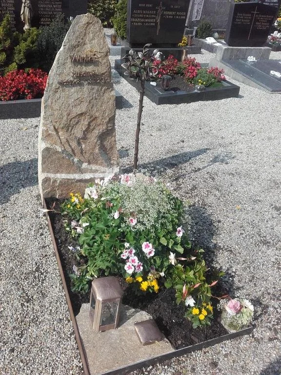 Ein Grabbild mit Blumen, einem Steinmonument, einer kleinen Laterne und einem kleinen Holzstuhl, in einem Friedhof.