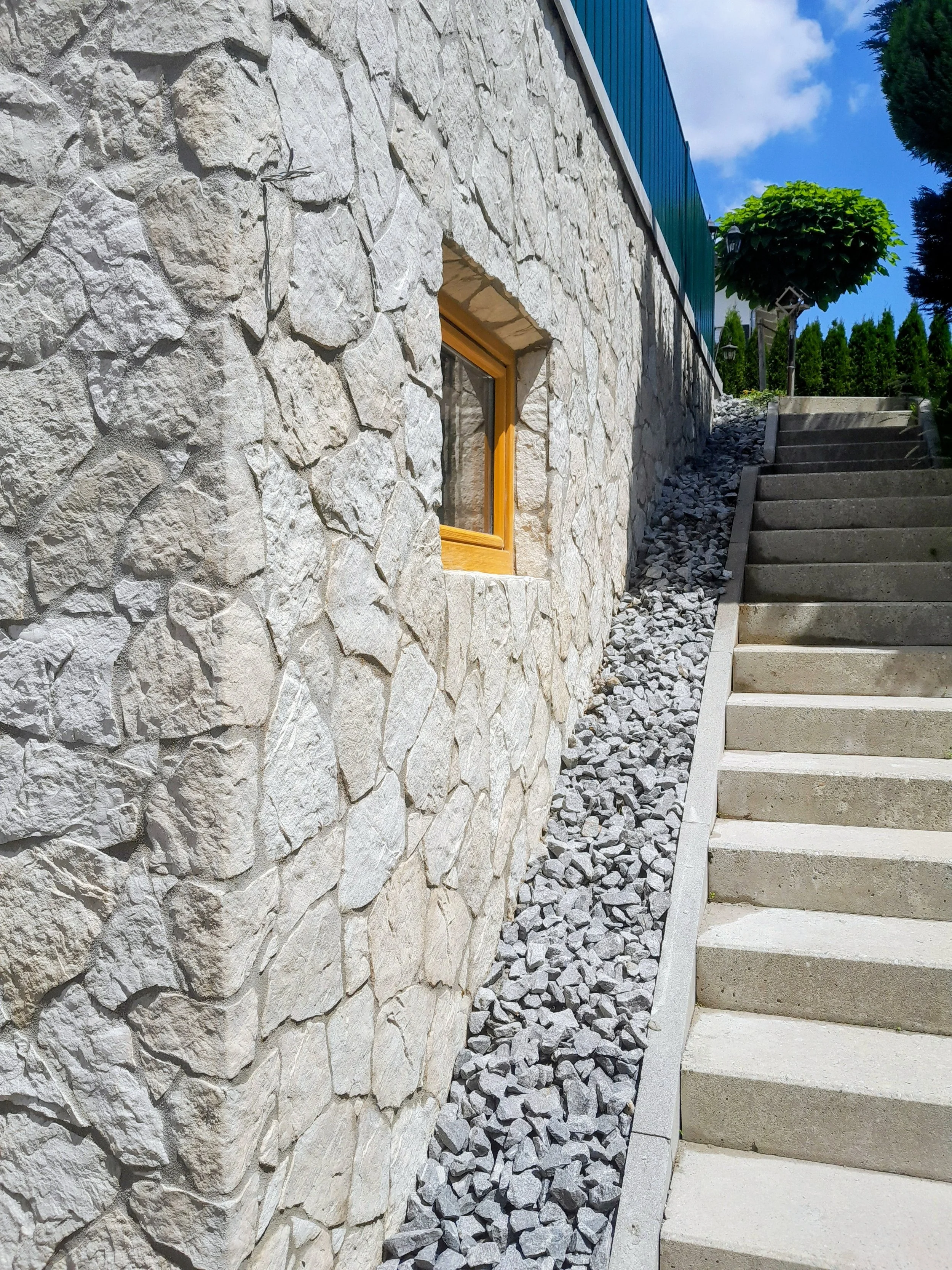 Außenwand eines Hauses mit Natursteinen, kleiner rechteckiger Fensterrahmen aus Holz, steile Treppe mit Betonstufen, Gräser und Büsche im Hintergrund, blauer Himmel mit Wolken.