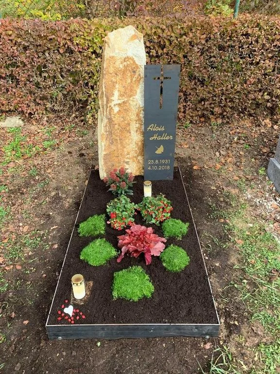 Ein Grab mit einem schwarzen Grabstein mit Name, Daten und einer kleinen Fliege, umgeben von Pflanzen und Kerzen.