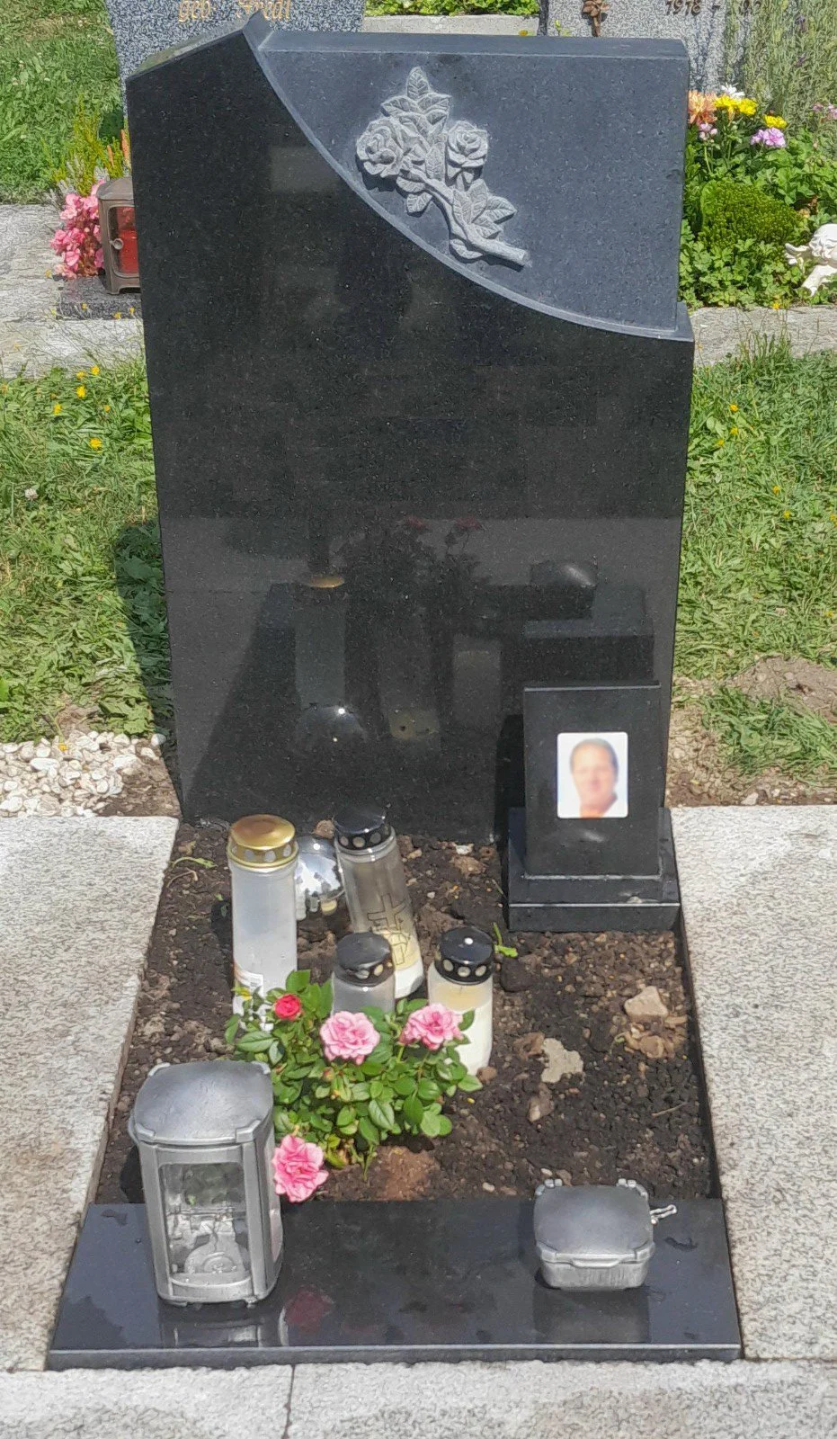 Schwarzer Grabstein mit Rosenrelief, Blumen, Kerzen und einem Foto des Verstorbenen auf einem Friedhof.