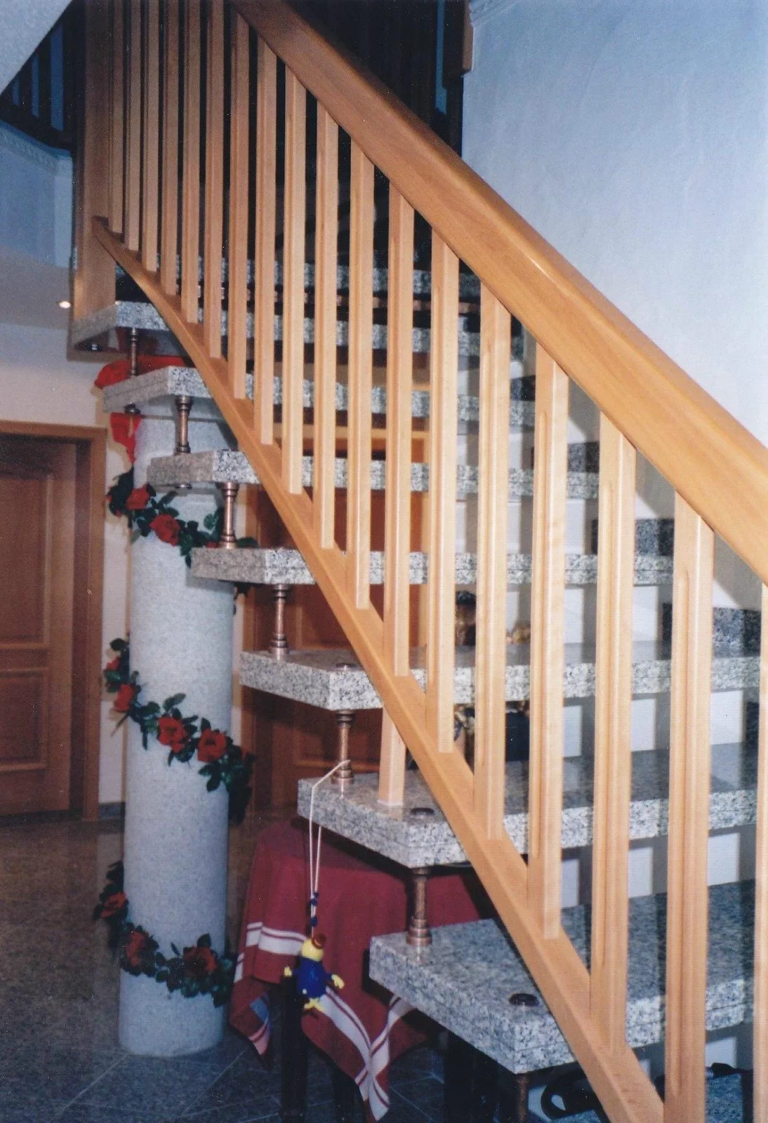 Innenaufnahme einer Treppe mit Granitstufen, Holzgeländer und einem dekorativen Pfeiler mit roten Blumen.