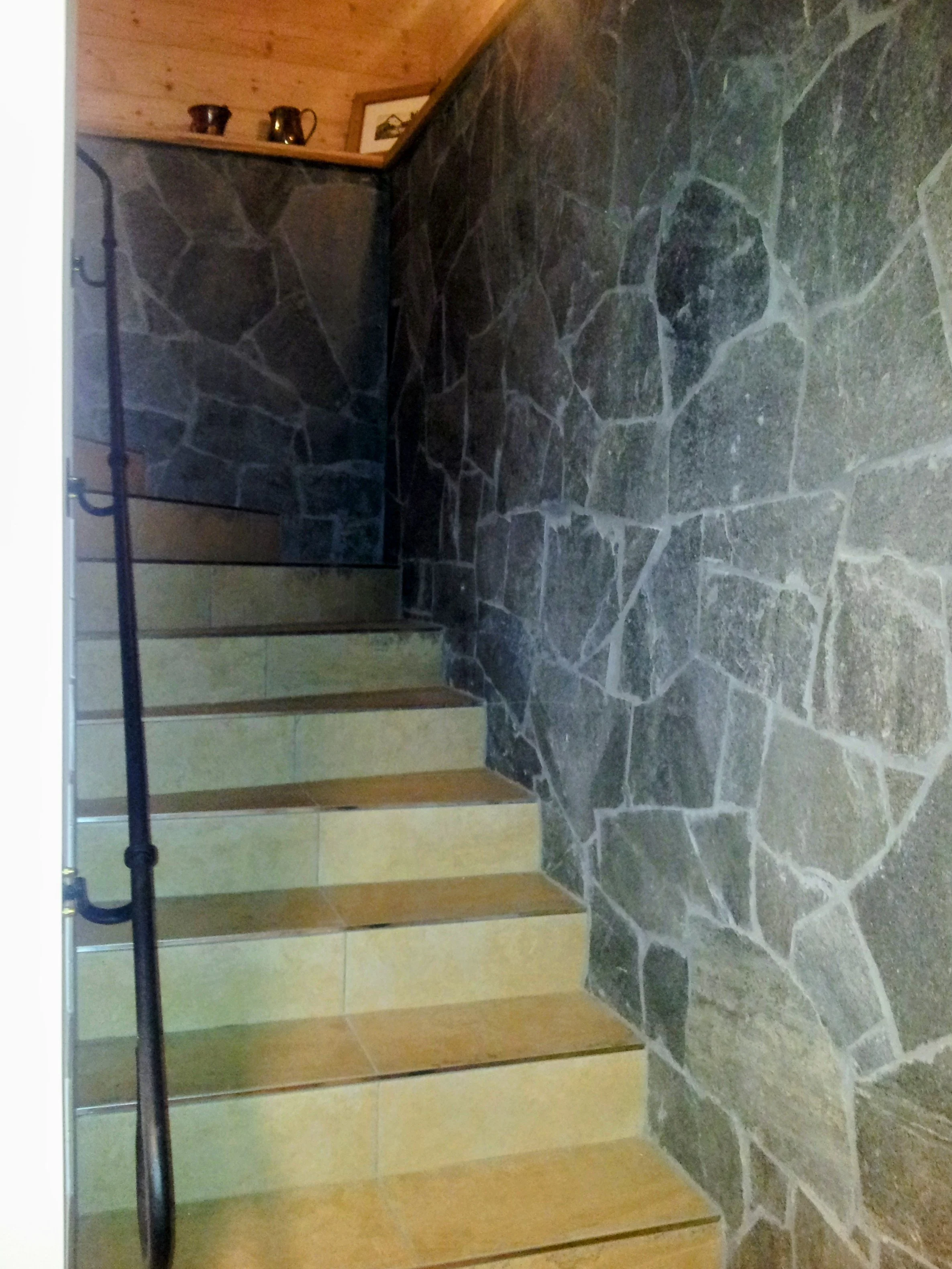 Innenansicht einer Treppe mit Holzstufen, links ein schwarzes Geländer, im Hintergrund eine Wand aus dunklen Steinen und eine Holzdecke, oben auf der Wand befinden sich drei kleine Kupferkrüge und ein Bildrahmen.