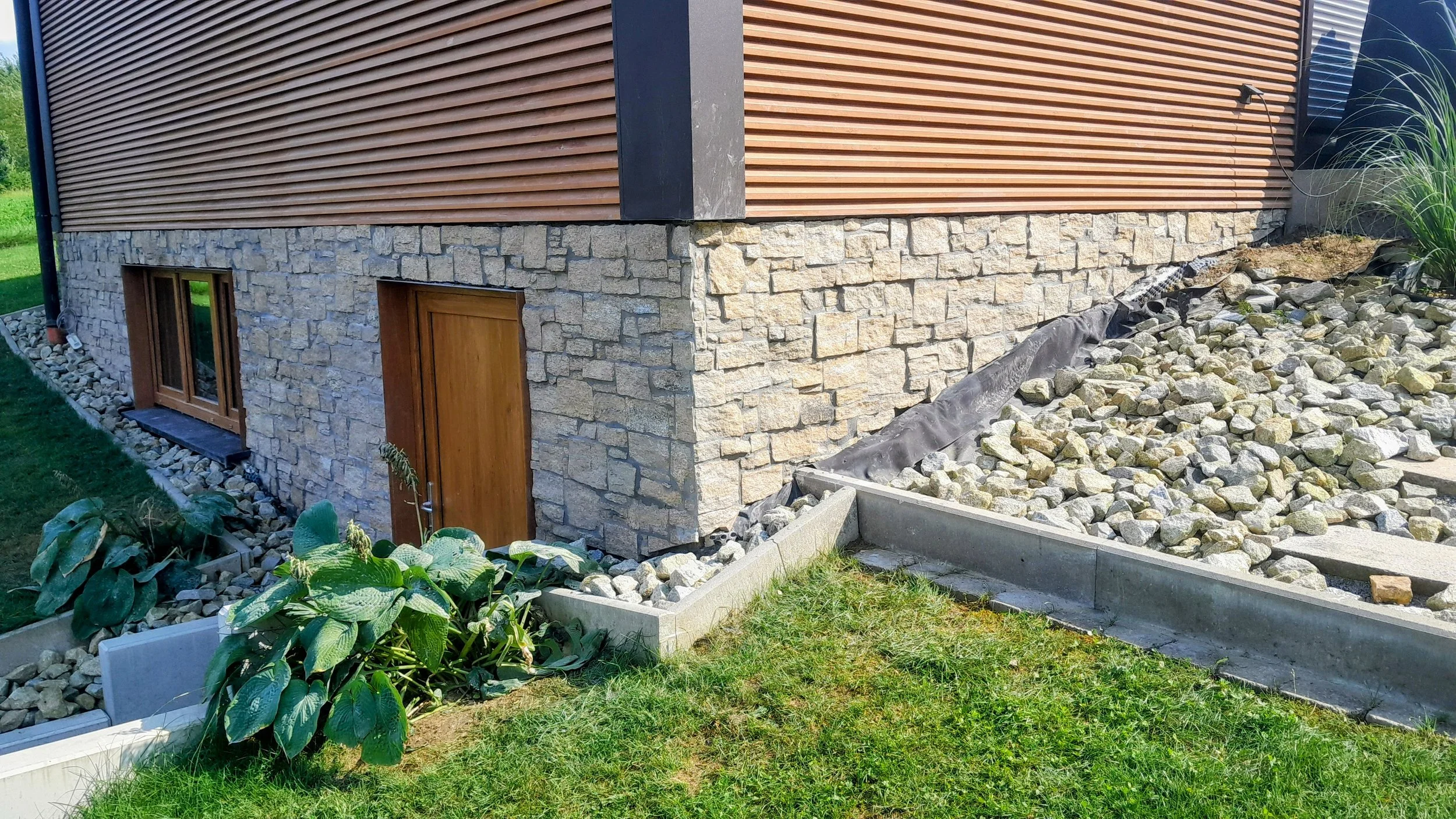 Teil eines Hauses mit Stein- und Holzfassade, Bauarbeiten im Gange, Steine und Bepflanzung im Vordergrund.