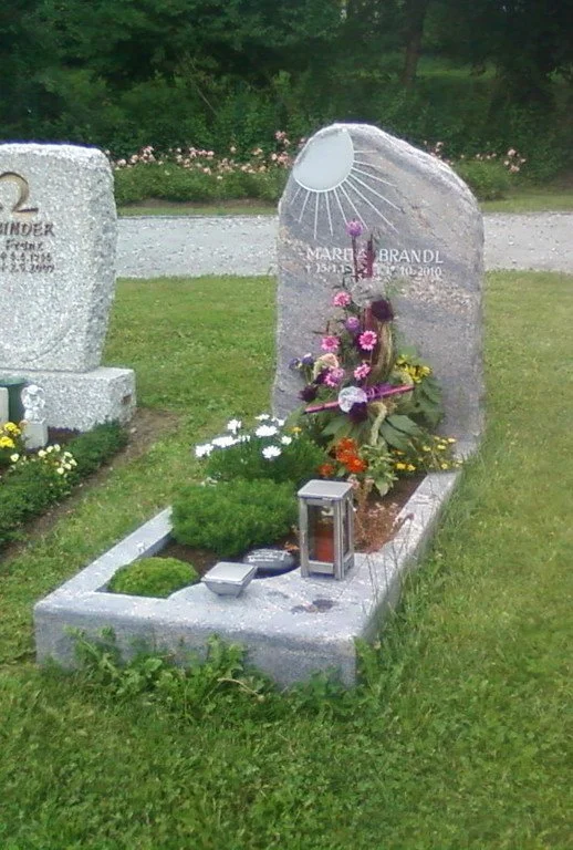 Ruhige Friedhofsszene mit liebevoll gestaltetem Einzelgrab. Moderner Grabstein aus Granit mit Sonnenmotiv, frische Blumen, Laterne und gepflegte Grabbepflanzung auf grüner Rasenfläche. Symbol für Erinnerung, Trost und würdige Grabgestaltung.