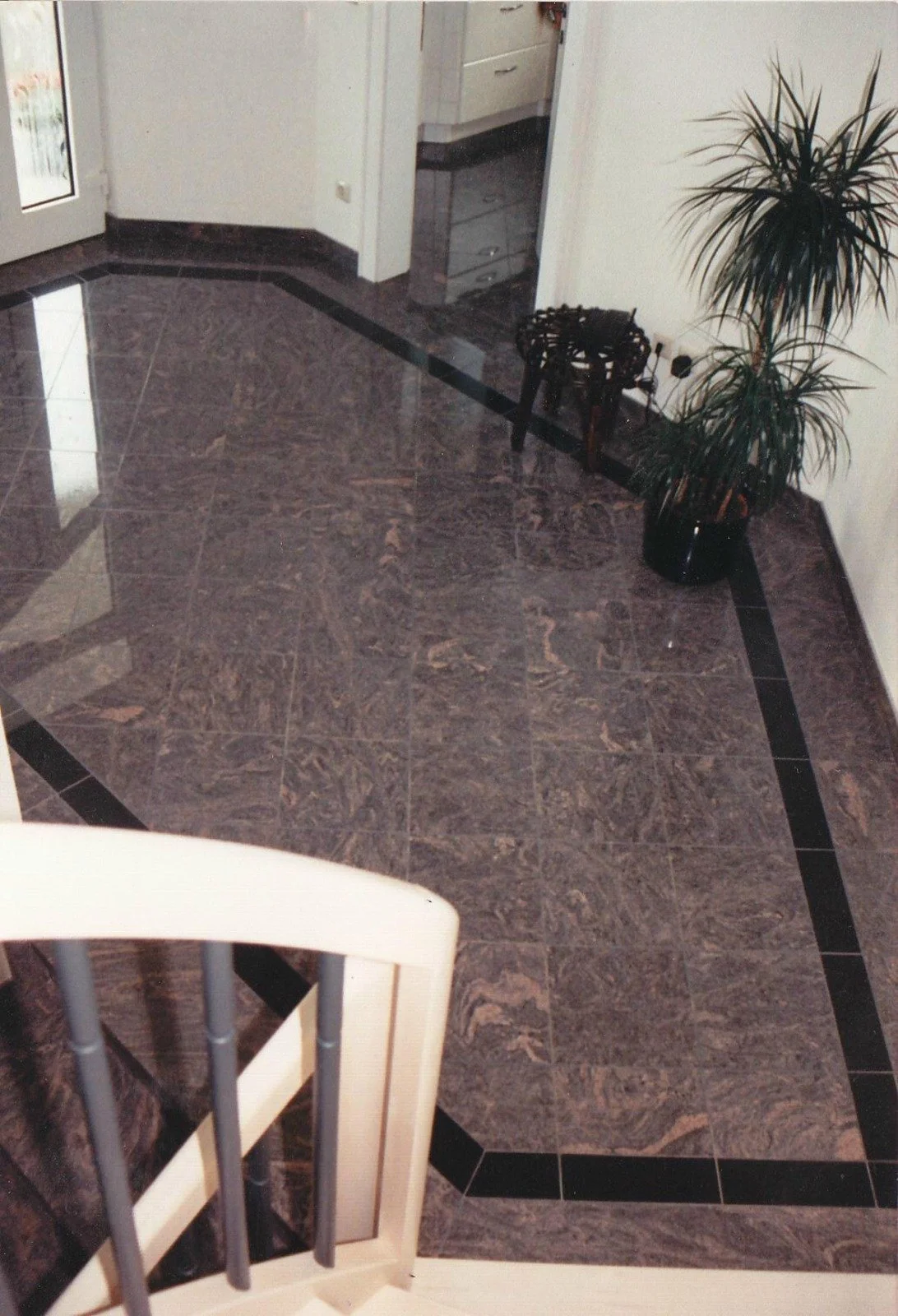 Bodenfliesen in einem Wohnraum mit dunklen Marmormustern, schwarzem Rand und weißen Wänden. Es sind ein Teil eines weißen Stuhls, eine Topfpflanze, ein kleiner Tisch und eine Tür sichtbar.